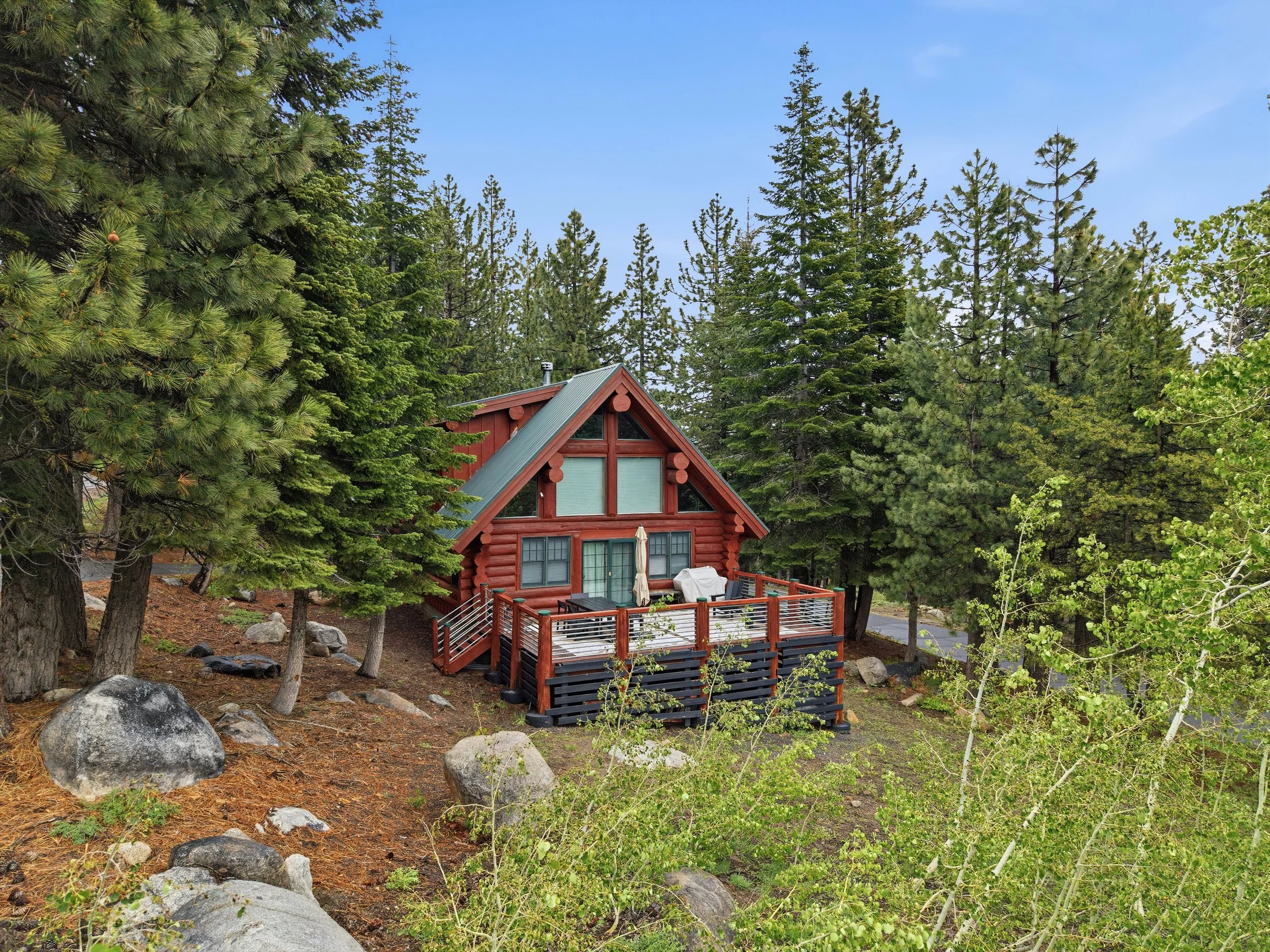 A red wooden cabin with a gabled roof and large windows, surrounded by tall pine trees and greenery, with a deck and outdoor furniture in the foreground and a clear blue sky overhead.