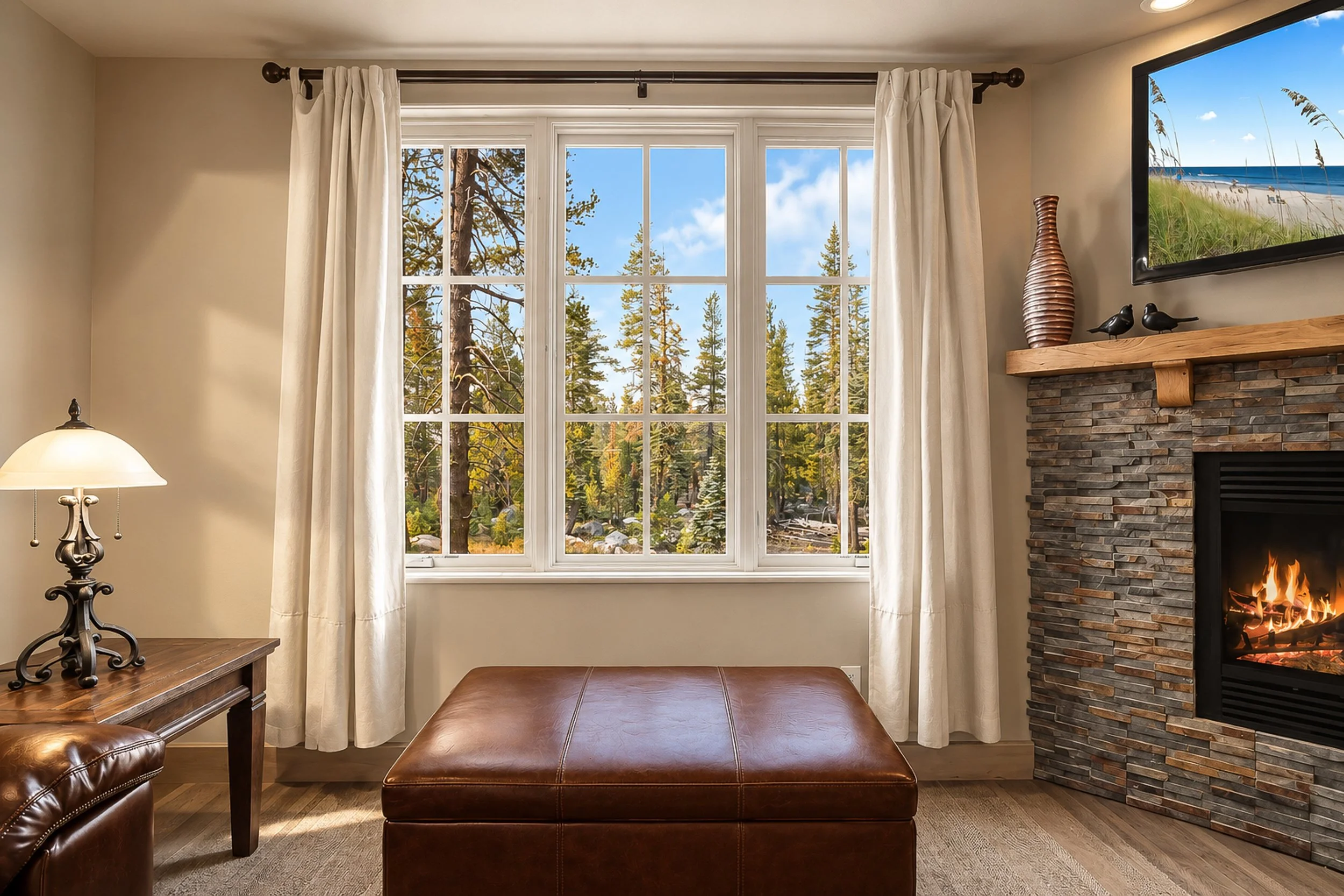 Living room with a large window showing trees outside, beige curtains, a fireplace with a stone surround and a TV mounted above, a table lamp on a side table, and leather furniture.