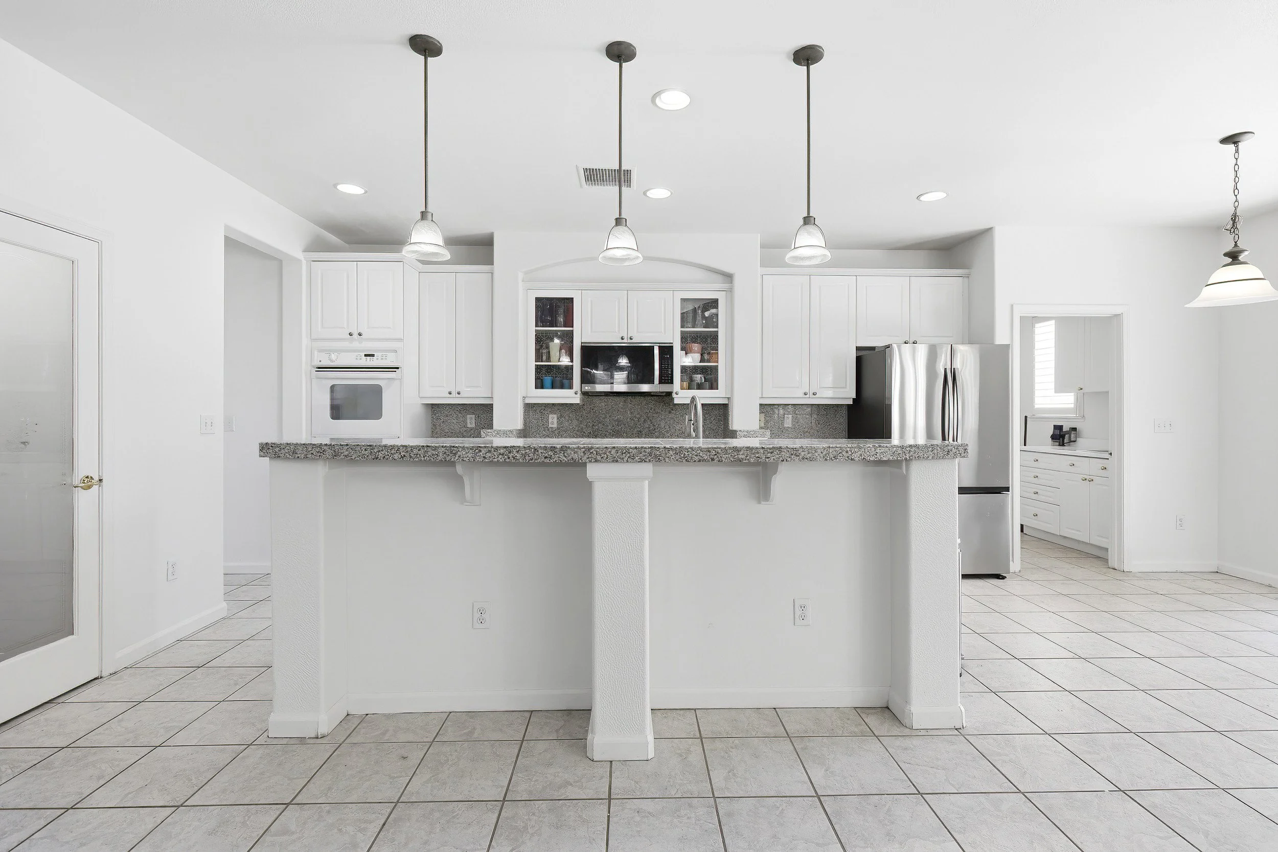A white kitchen with granite countertops, white cabinets, and stainless steel appliances. There are three pendant lights hanging above the counter and a pot rack lighting fixture on the right. The floor is tiled with large white tiles.