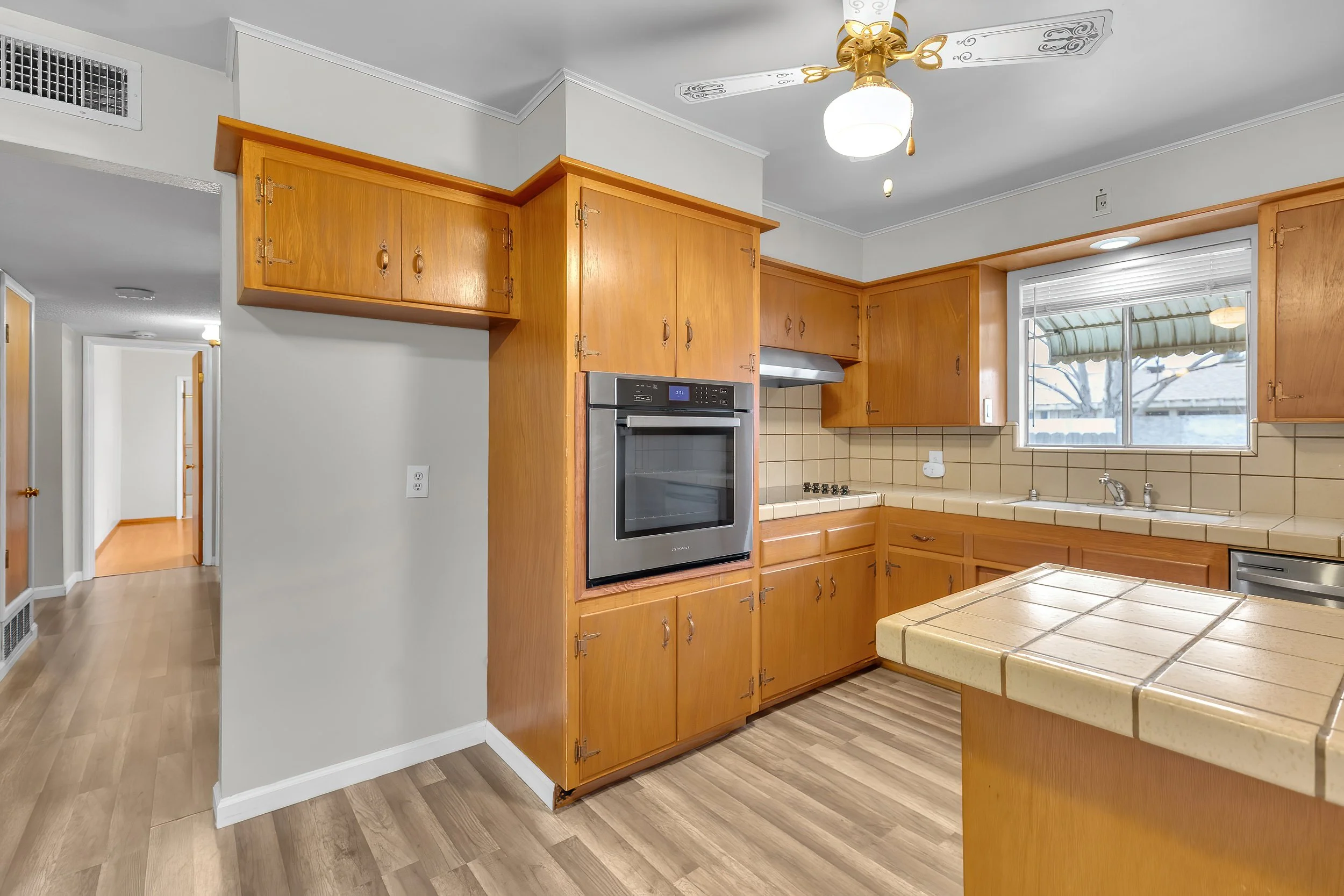 Kitchen with wooden cabinets, a stove, and a window overlooking an outdoor scene with trees. Ceiling fan with light. Light-colored walls and wood flooring.