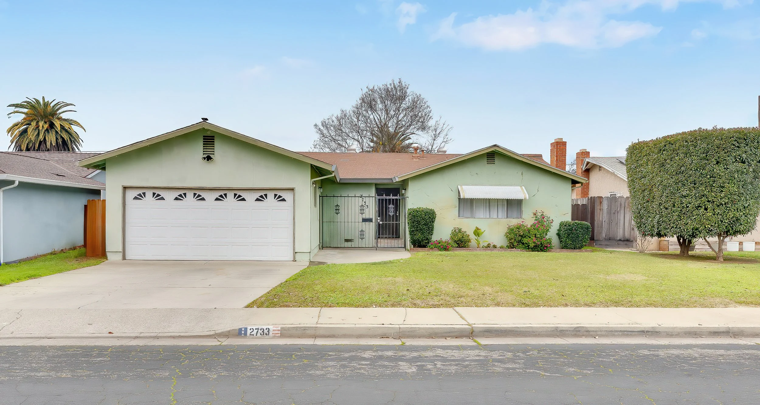 A single-story house painted light green with a white garage door and a black metal gate in front. There are small shrubs and flowers in the yard, a tall hedge on the right, and a large tree behind the house. The house number 2733 is on the sidewalk 