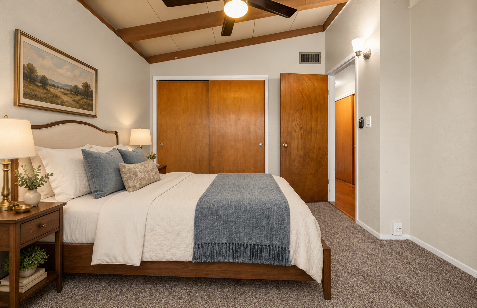 A cozy bedroom with a large bed covered in white bedding, blue pillows, and a gray throw blanket. Beside the bed are wooden nightstands with lamps and potted plants. There is a landscape painting above the bed, wooden closet doors, and a doorway lead