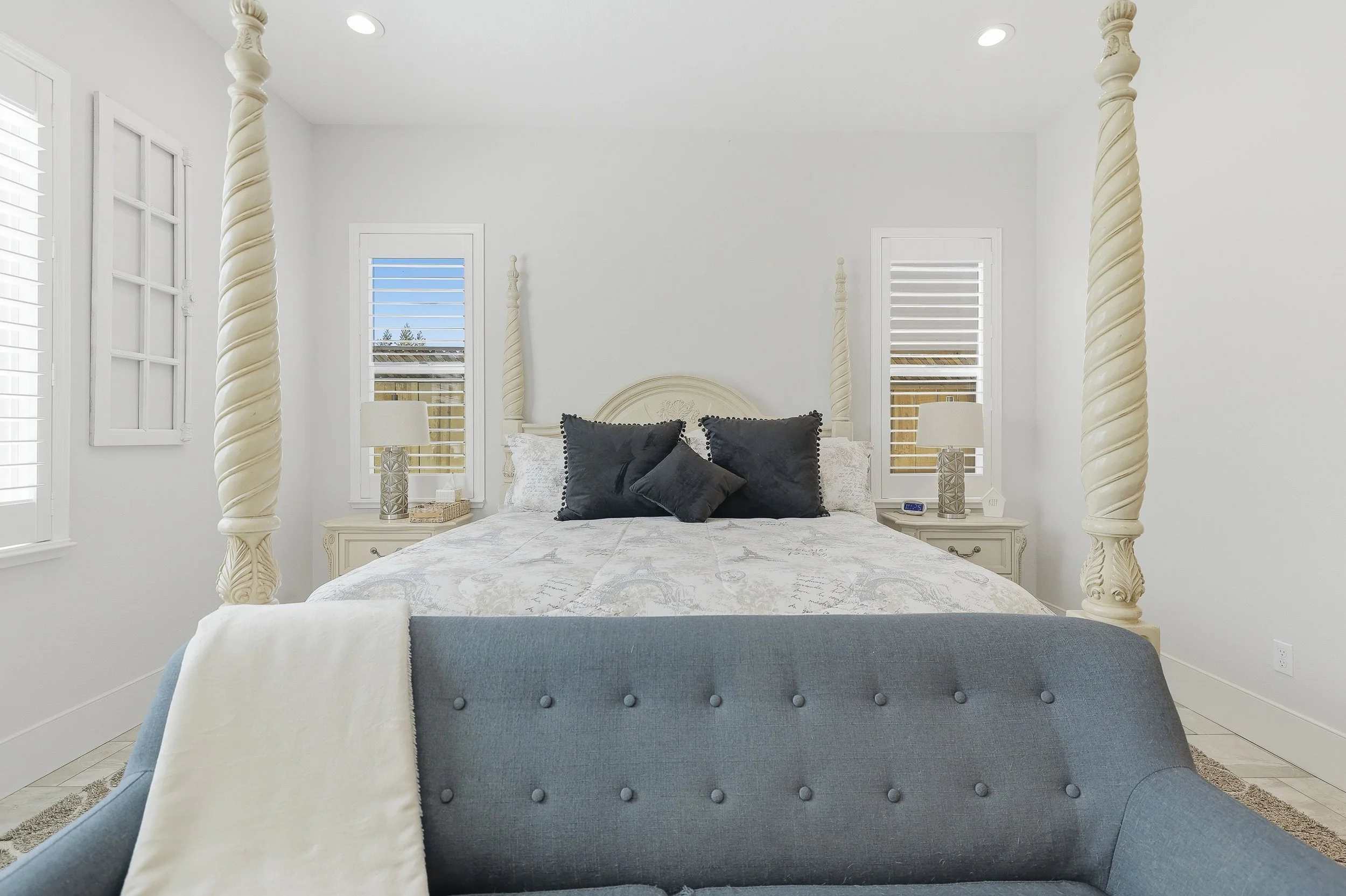 A bedroom with a four-poster bed with black pillows, white bedding, and two matching lamps on bedside tables, in a bright room with white walls and windows.
