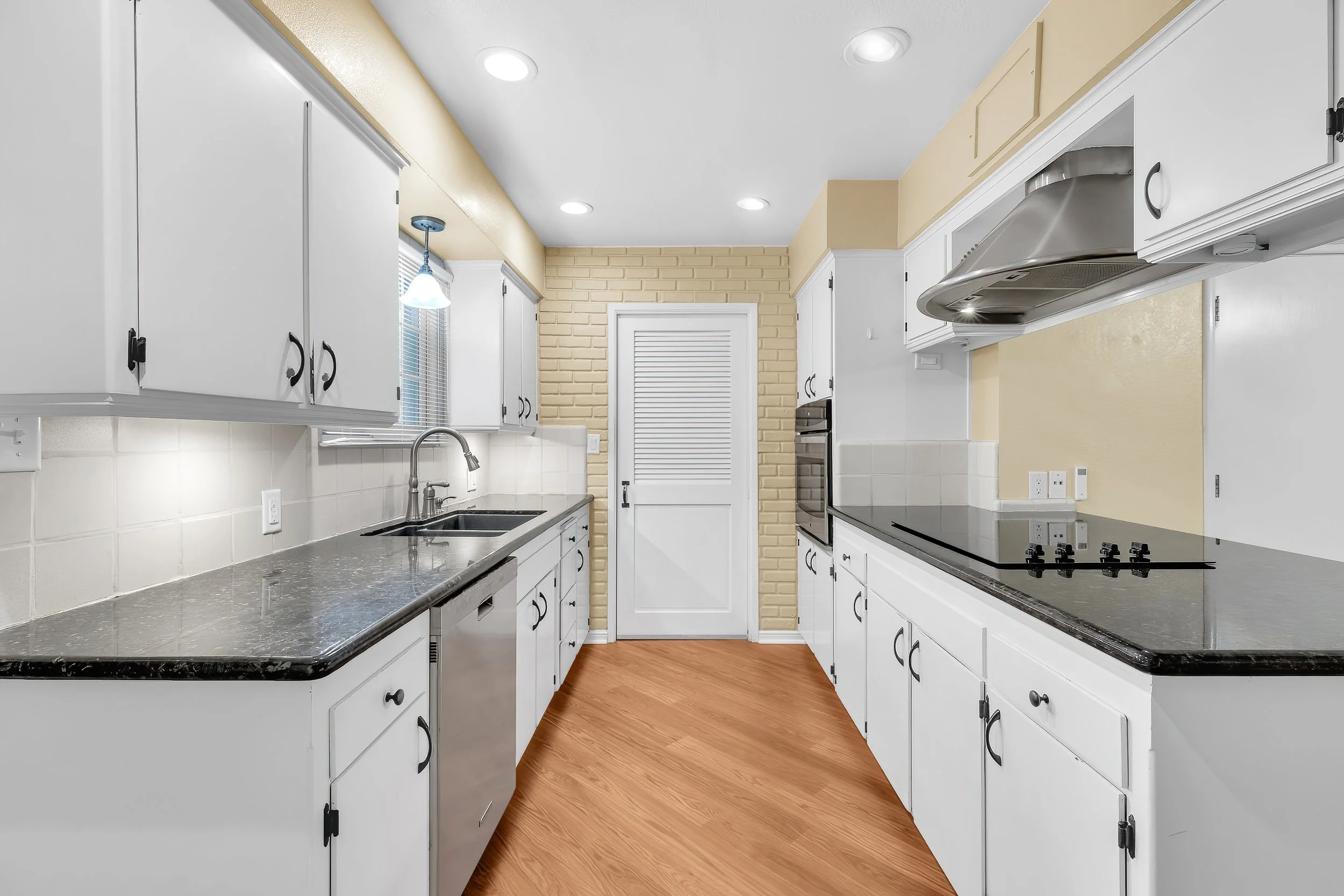 A kitchen with white cabinets, black handles, a gray countertop, a double sink, and a window. There is a stove with black burners, a range hood, and a brick wall near a white door with vent fins. Recessed ceiling lights and wood flooring are also pre