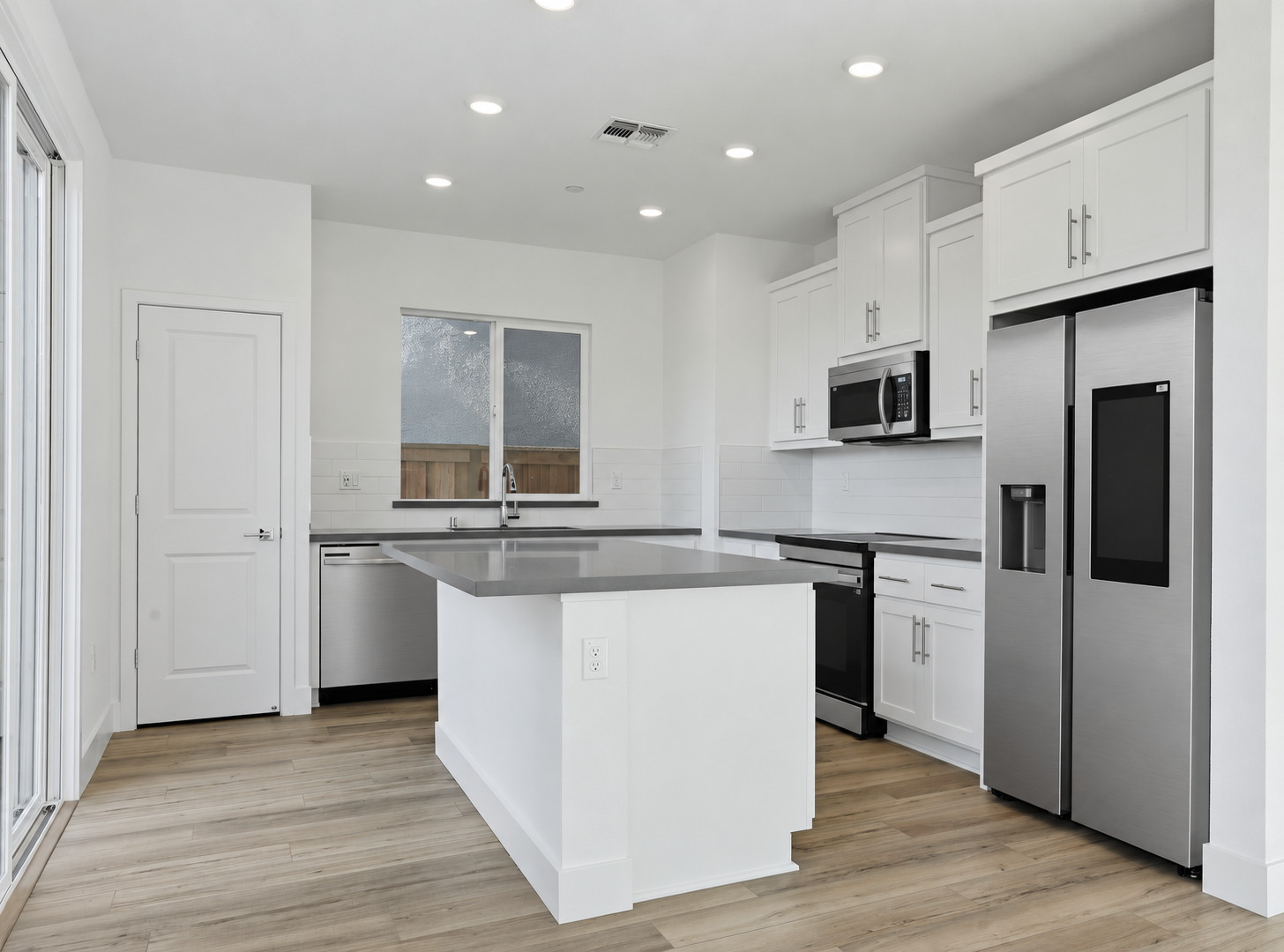Modern kitchen with white cabinets, stainless steel appliances, black countertops, island with gray countertop, wood flooring, large window, and recessed ceiling lights.
