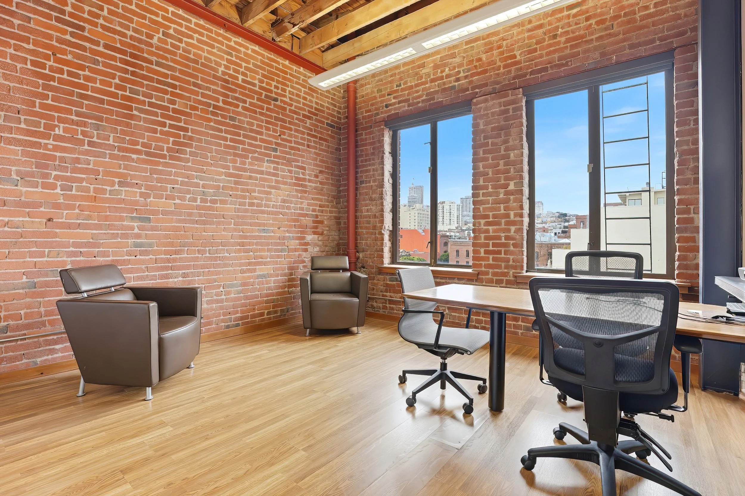 Modern office with exposed brick walls, wooden floor, large windows showing cityscape, and office furniture including chairs and a desk.