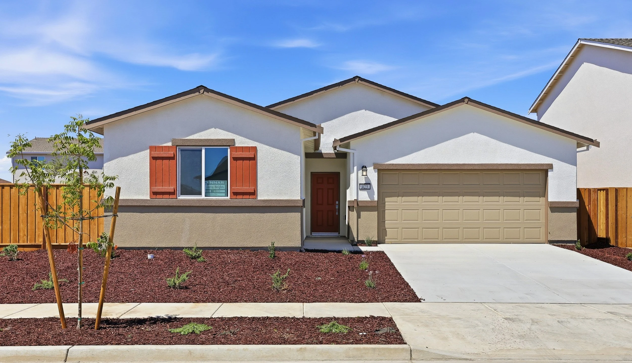 Front view of a modern two-story house with a beige garage door, red front door, white exterior walls, and a small window with red shutters. There is a small front yard with newly planted greenery and red mulch, and a gravel sidewalk in front.