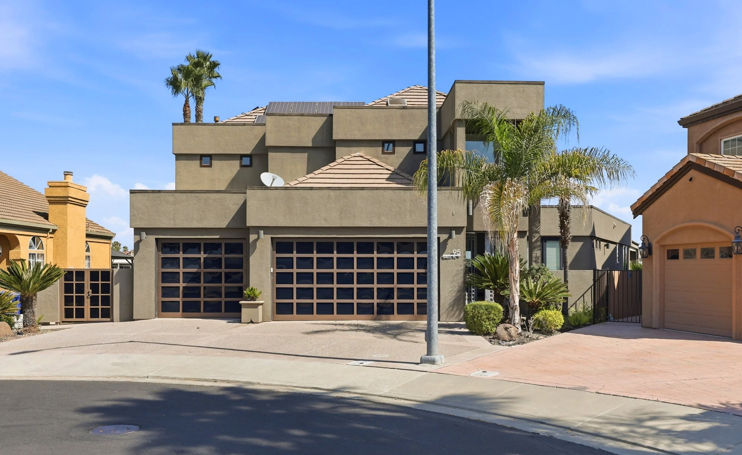 A modern multi-story house with beige and gray exterior walls, a tiled roof, and palm trees in the front yard, located in a sunny neighborhood with a clear blue sky.