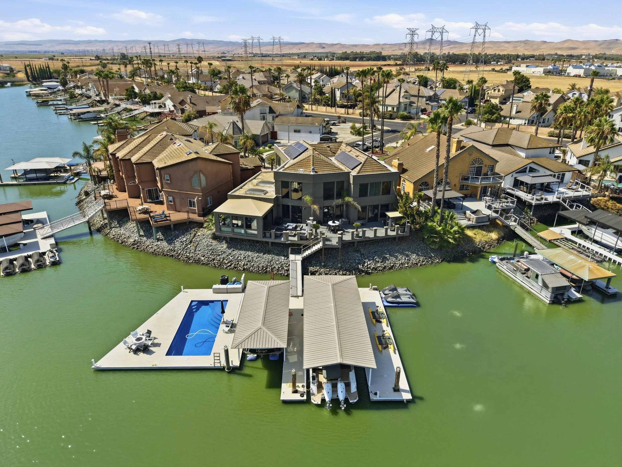 Aerial view of a waterfront residential area with houses, docks, and boats along a greenish waterway, with hills and open fields in the background.