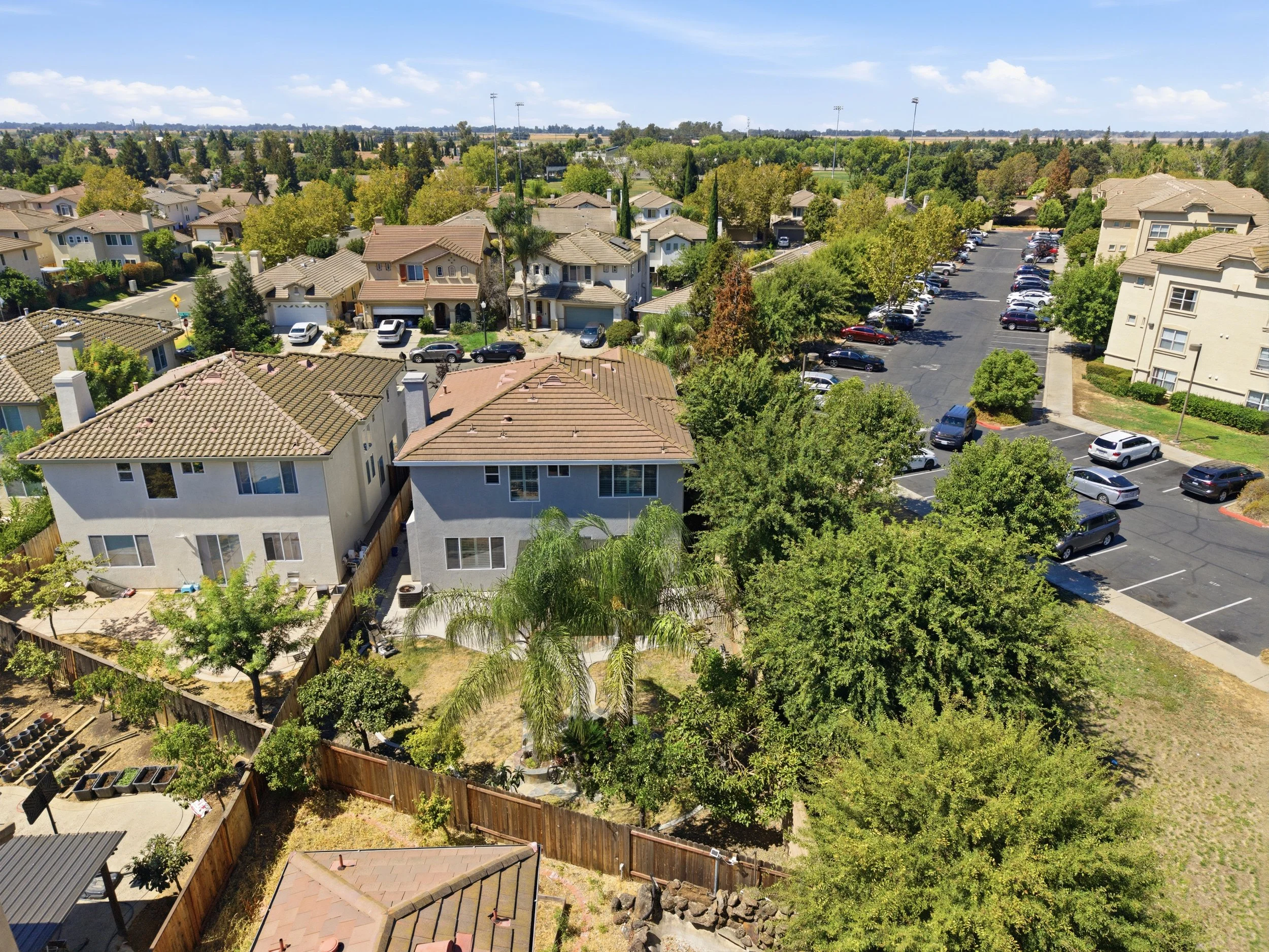 Aerial view of a suburban neighborhood with houses, trees, a parking lot, and a grassy area under a partly cloudy sky.