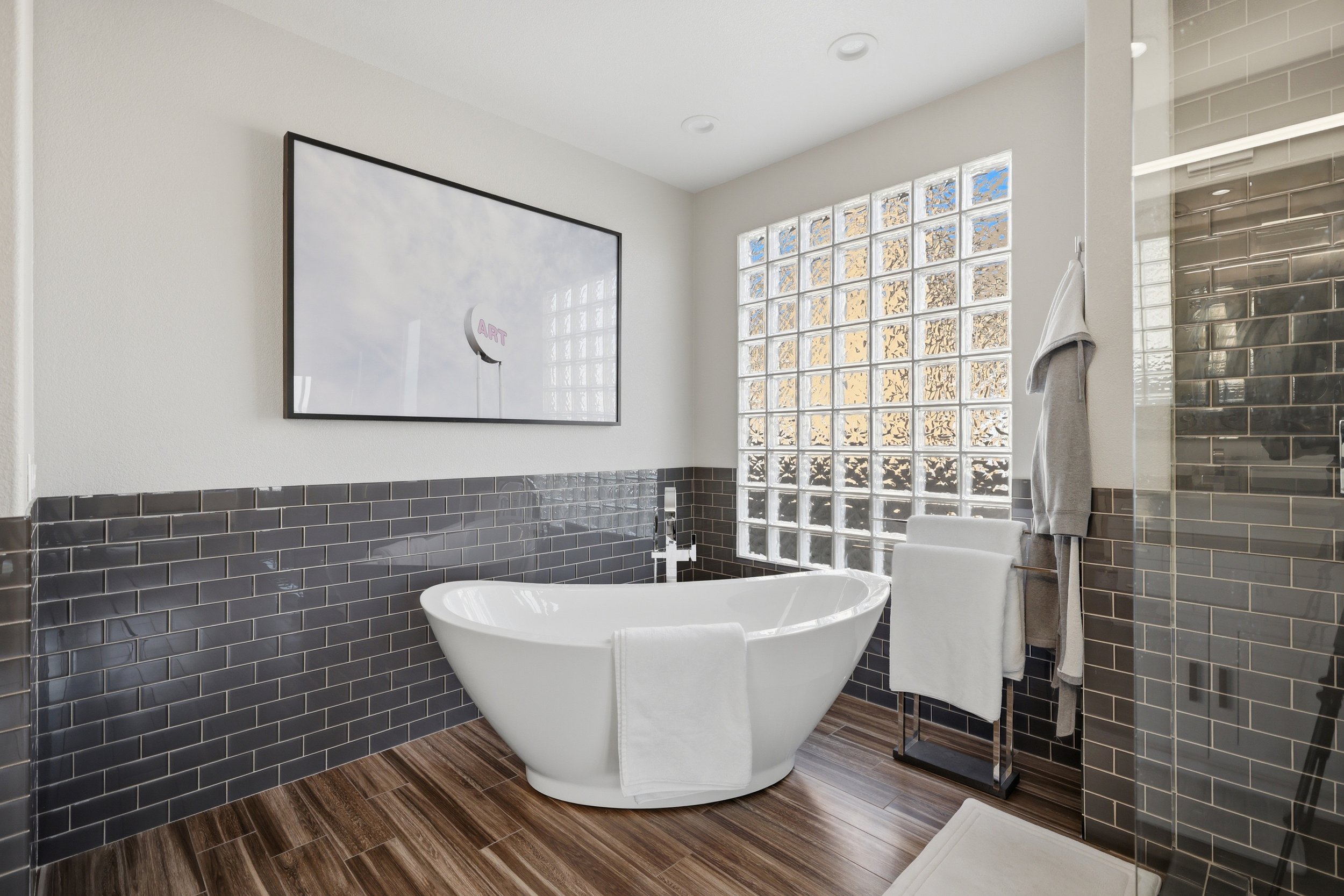 Modern bathroom with a freestanding bathtub, gray tiled wall, glass block window, dark wood floor, gray towel hanging on a stand, and a large framed art piece on the white wall.