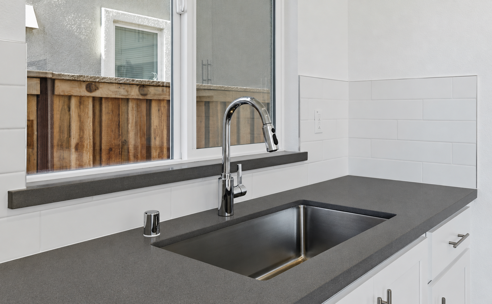 Modern kitchen countertop with a stainless steel sink, chrome faucet, and a window overlooking a wooden fence.