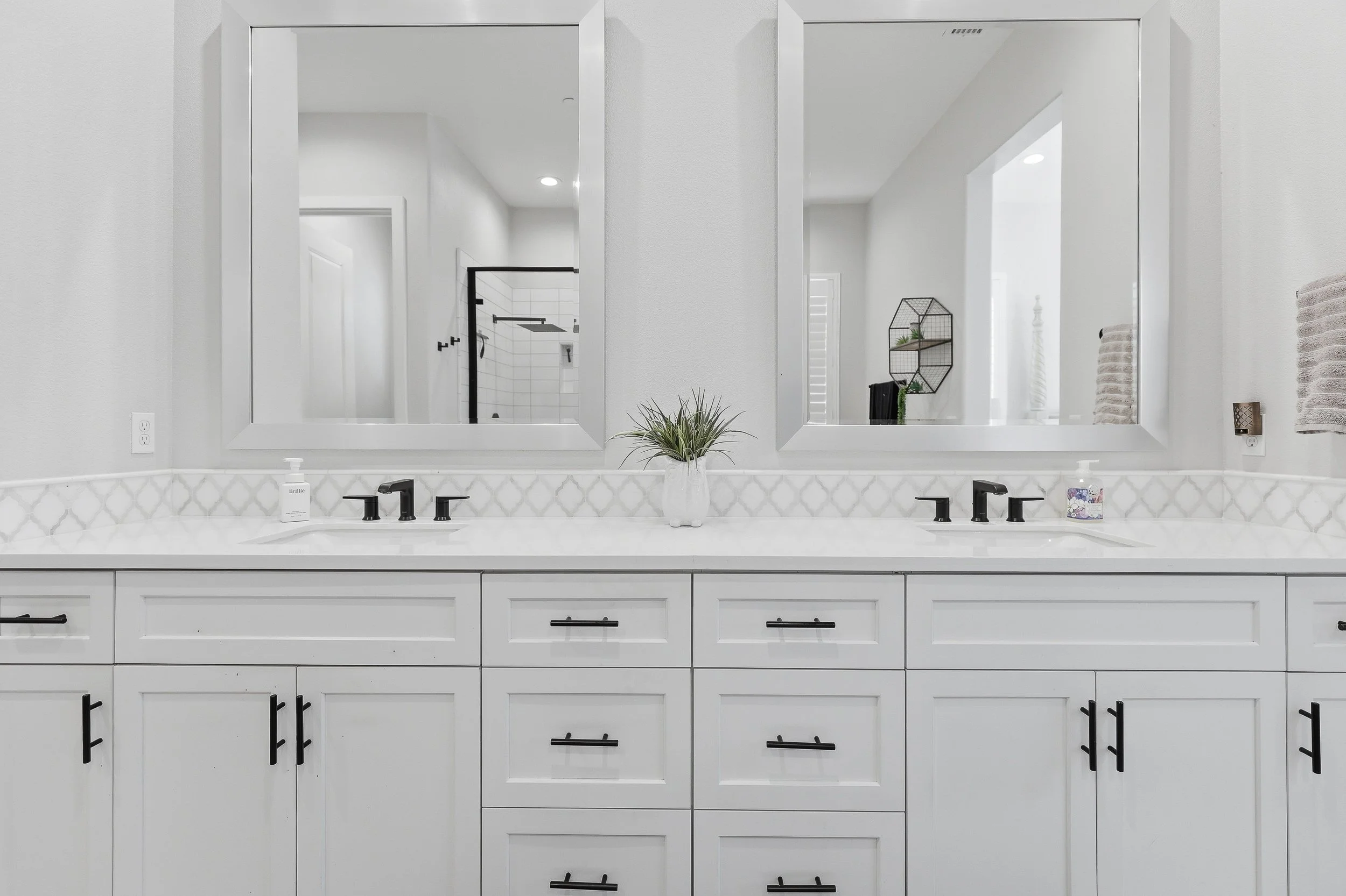 A bathroom vanity with two sinks, mirrors, and black faucets, decorated with a small plant and a candle, with a view into a shower with a black frame and white tile walls.