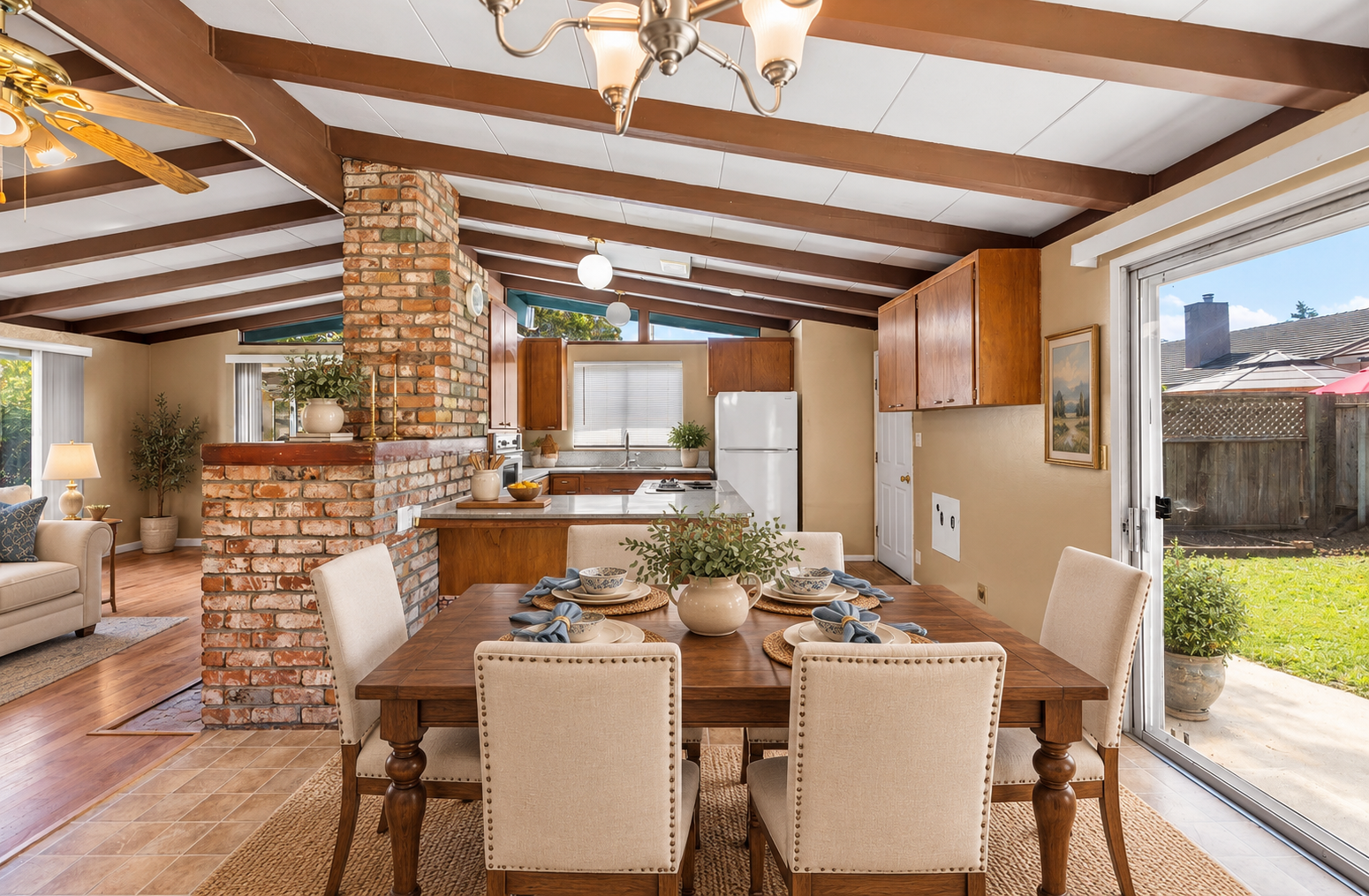 Open-concept kitchen and dining area with brick support wall, wooden beams, beige chairs around a wooden table with a floral centerpiece, natural light from sliding glass door showing backyard.
