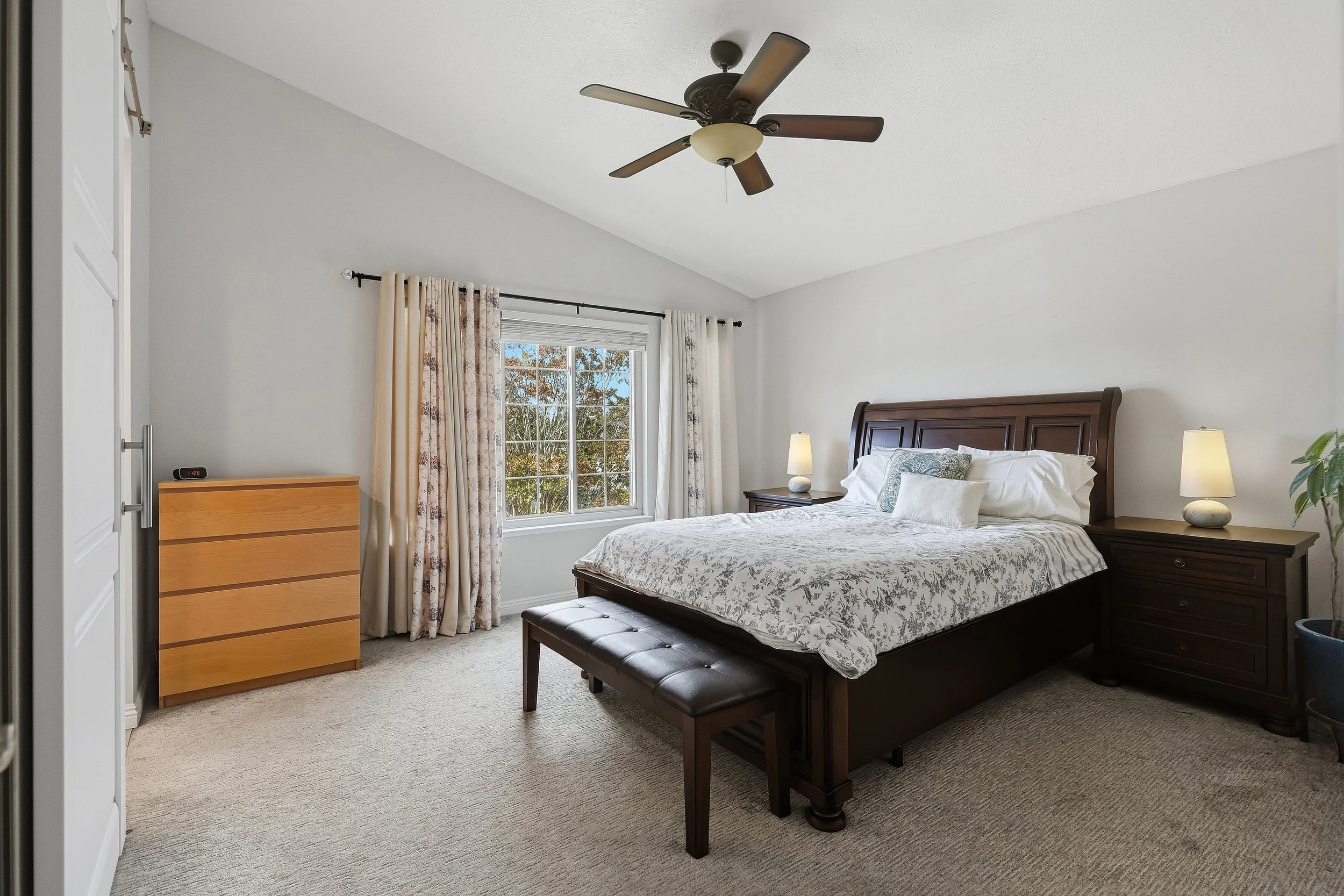Bedroom with dark wood bed, ceiling fan, and windows overlooking greenery.