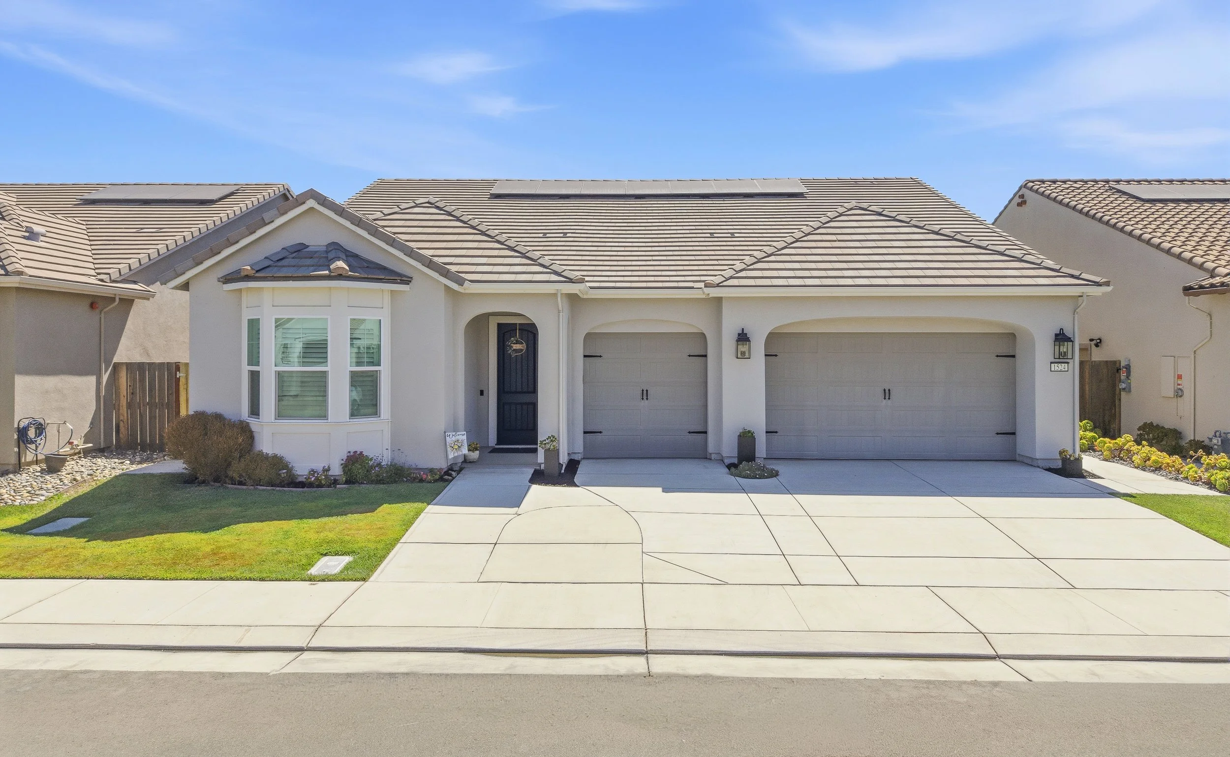 Front view of a modern, white house with a grey tiled roof, two garage doors, a black front door, and a green lawn with small bushes, under a blue sky.