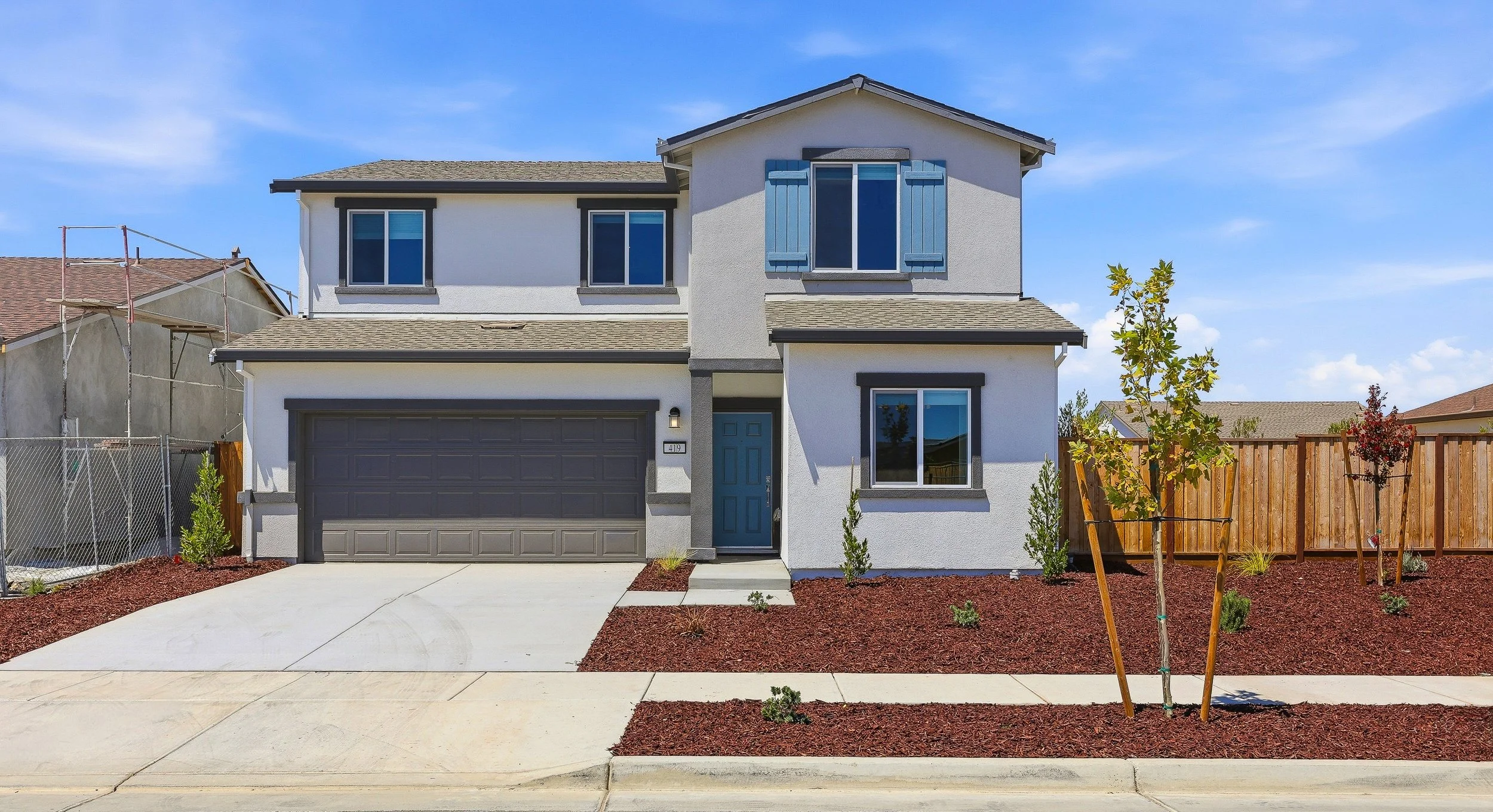 A modern two-story house with a light-colored exterior, blue shutters, and a dark gray garage door, set against a partly cloudy sky, with a small front yard landscaped with mulched planting beds and young trees.