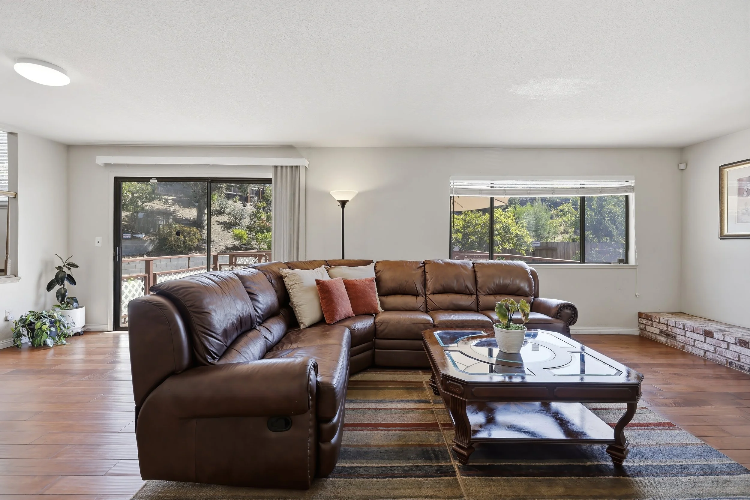 Living room with a brown leather sectional sofa, a wooden coffee table with decorative inlay, large windows with blinds, a potted plant, a standing lamp, and a brick fireplace.