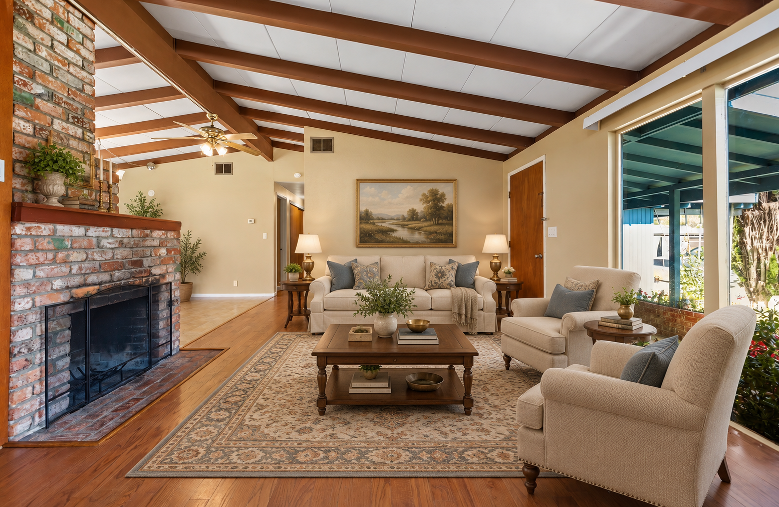Living room with a brick fireplace, beige sofa, two beige armchairs, dark wood coffee table, wall art, and large window facing a blue porch roof.