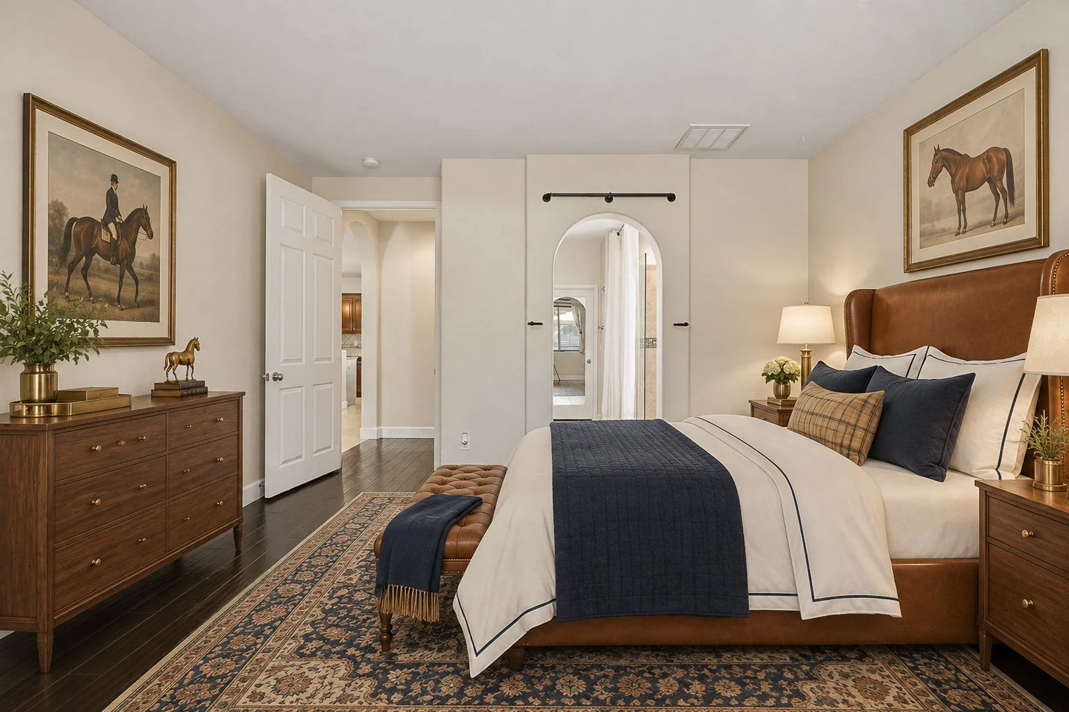 A cozy bedroom with a large bed featuring white and navy bedding, wooden nightstands with lamps, and framed equestrian art on the walls. There is a wooden dresser with decorative items and a tufted bench at the foot of the bed. The room has hardwood 