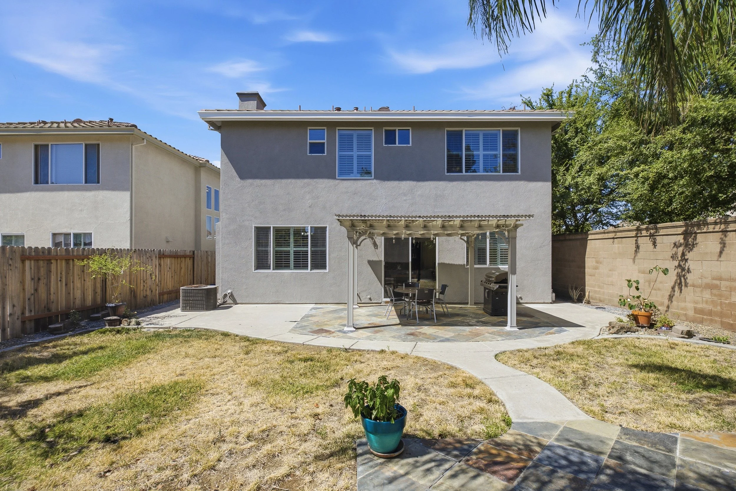 Backyard patio with a covered seating area, a barbecue grill, potted plants, a wooden fence, and part of a neighboring house with gray walls and several windows. There is a grassy lawn with a curved concrete walkway.
