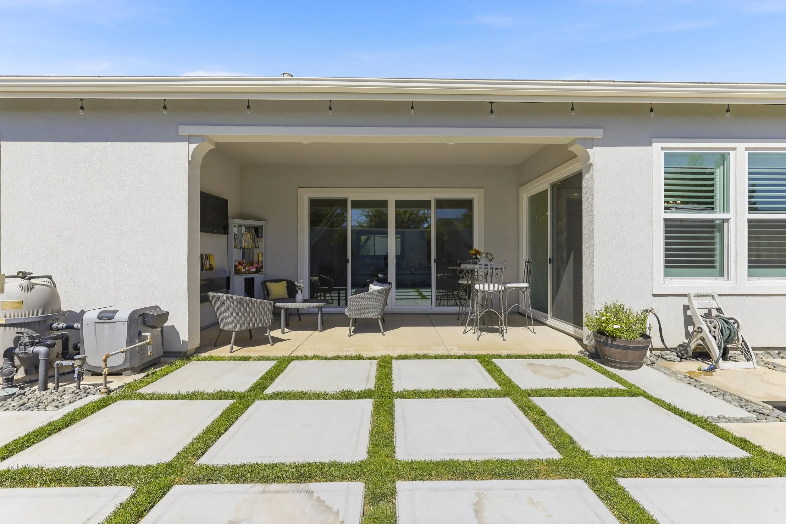 Backyard with patio furniture, potted plants, a grill, sliding glass doors leading into the house, and a lawn with concrete stepping stones.