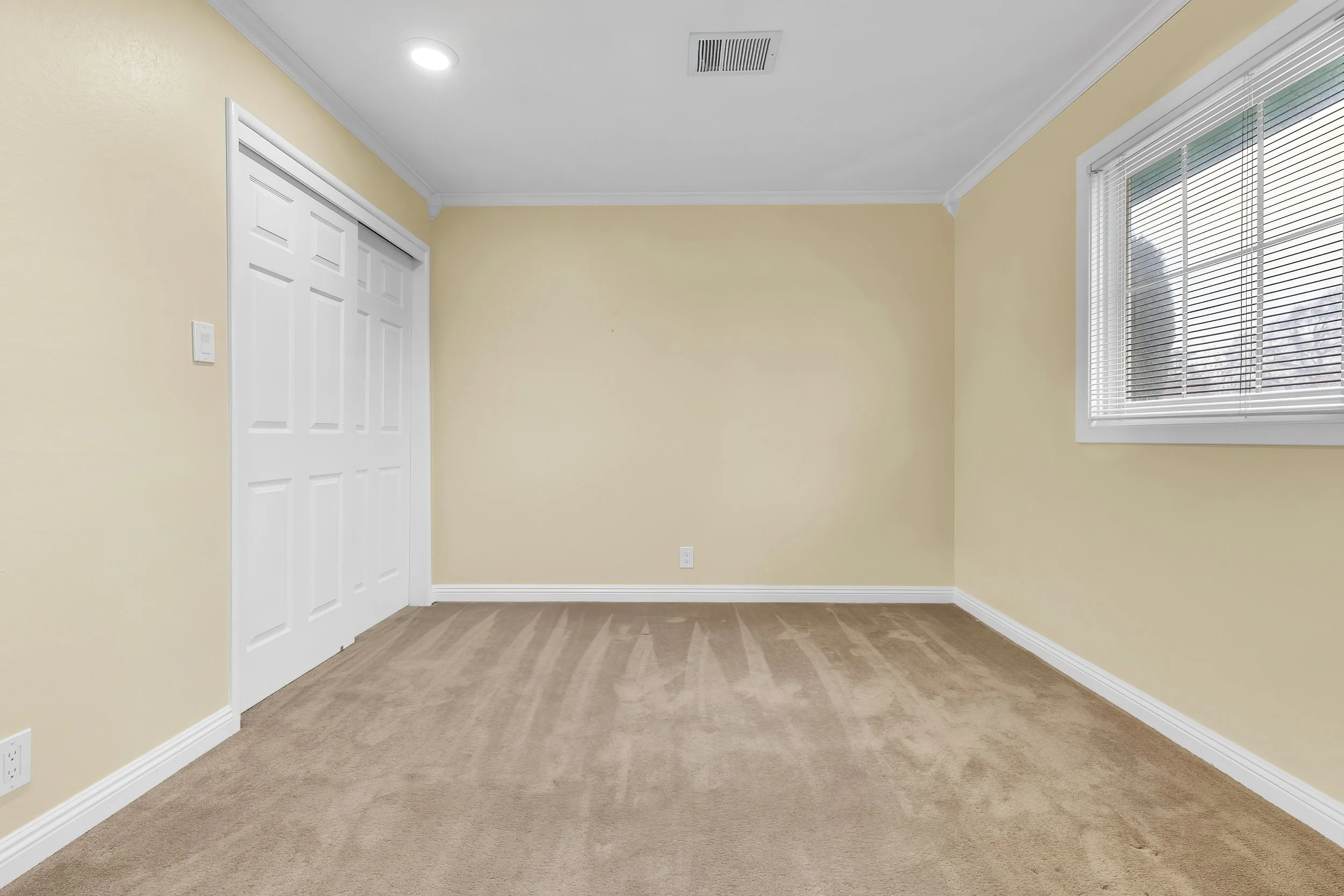 Empty room with beige walls, white trim, window with blinds, closet doors, ceiling light, and air vent.