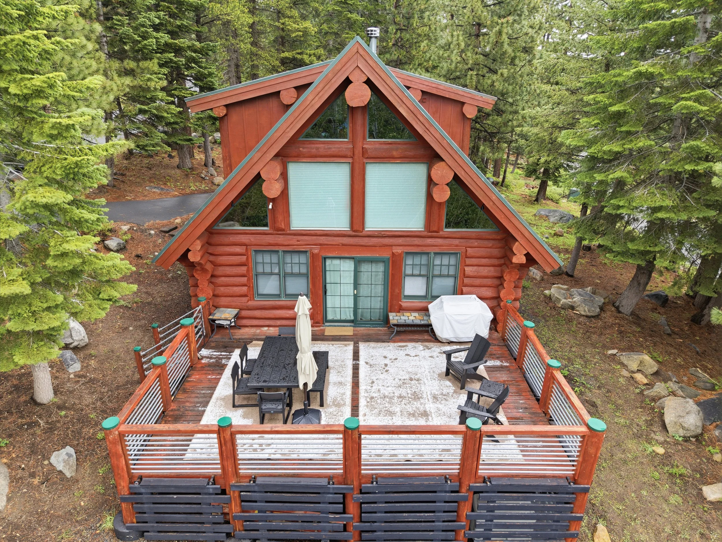 Aerial view of a log cabin with a steep A-frame roof surrounded by tall evergreen trees. The cabin has large triangular windows and a wooden deck with outdoor furniture including a table with an umbrella and several chairs. There is also a grill cove