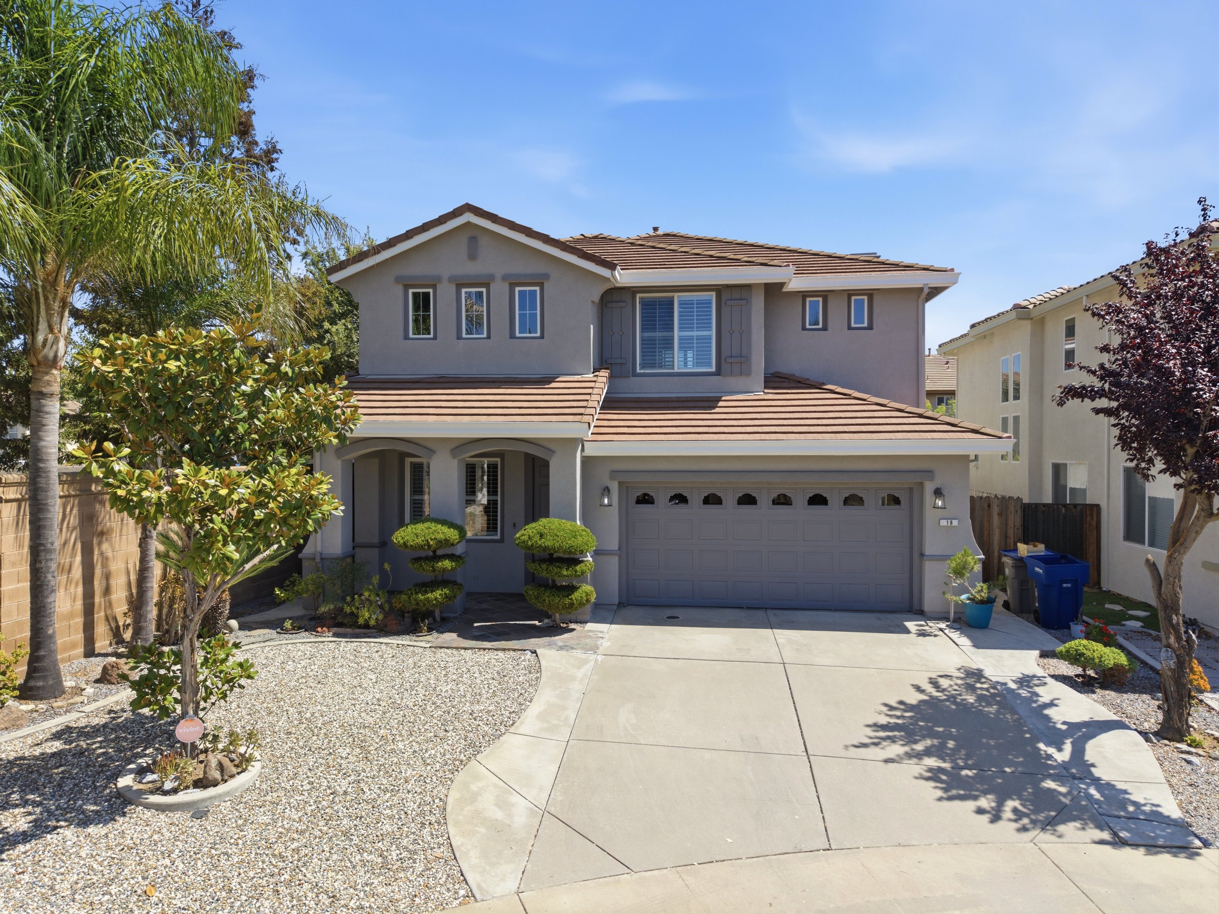 Front yard with a concrete driveway, trees, bushes, and a two-story house with a garage, under a clear blue sky.