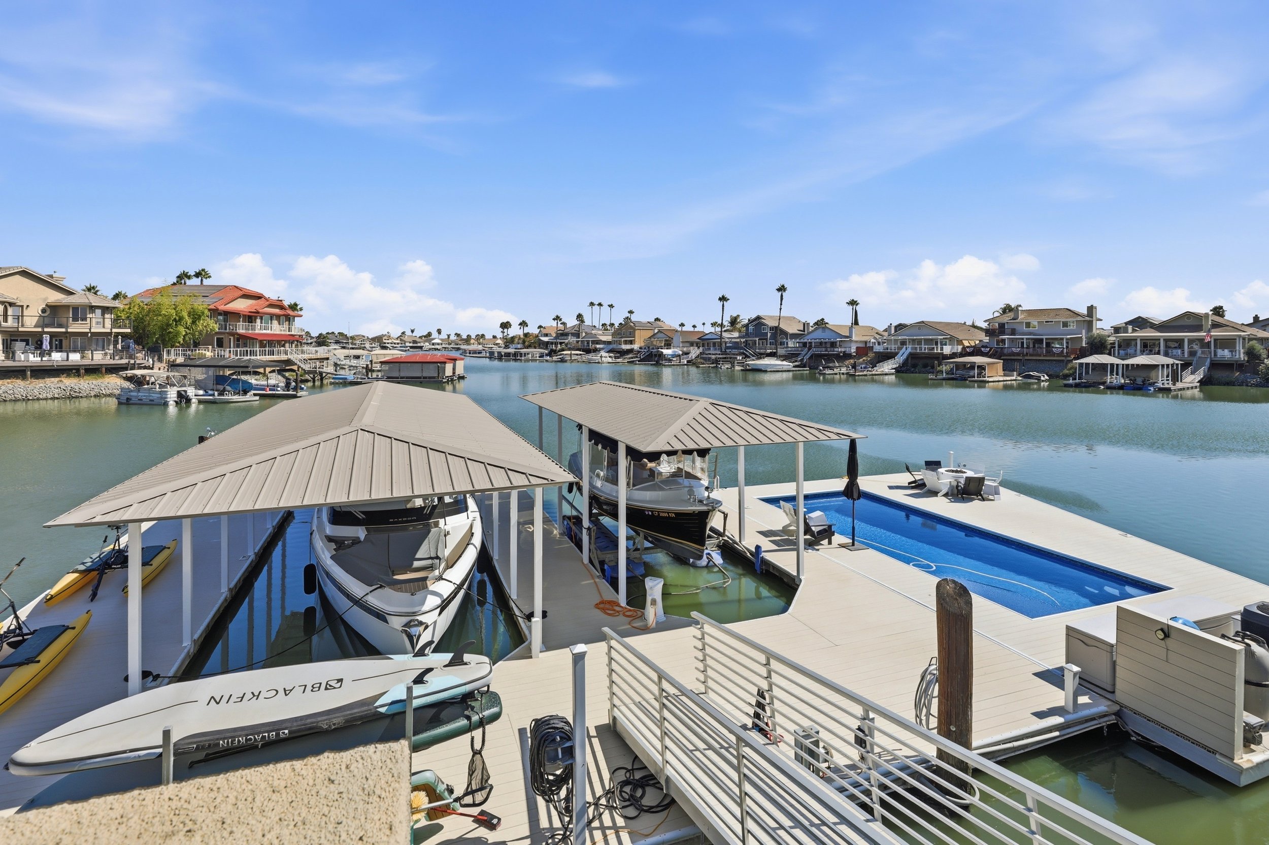 A waterfront scene featuring boats docked at a marina under covered slips, with a row of houses along a canal and palm trees in the background.