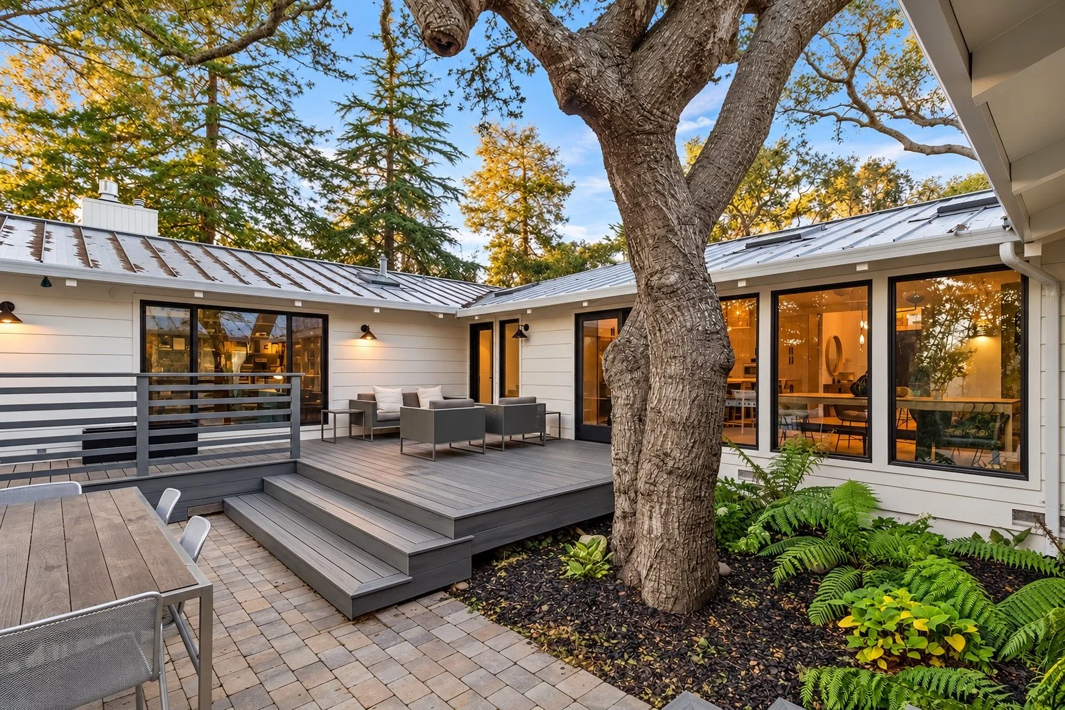 Carmel courtyard with a gray deck, outdoor seating, a mature tree, and illuminated windows.