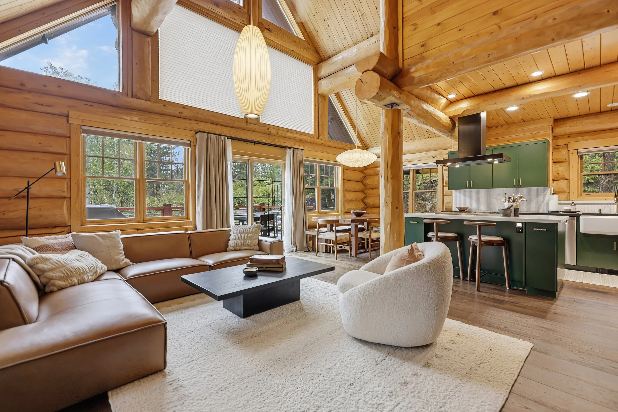 Interior of a log cabin-style living room with large windows, wooden beams, and a high vaulted ceiling. The room features a beige sectional sofa, a white armchair, a dark rectangular coffee table, and a cozy white rug. In the background, a dining are