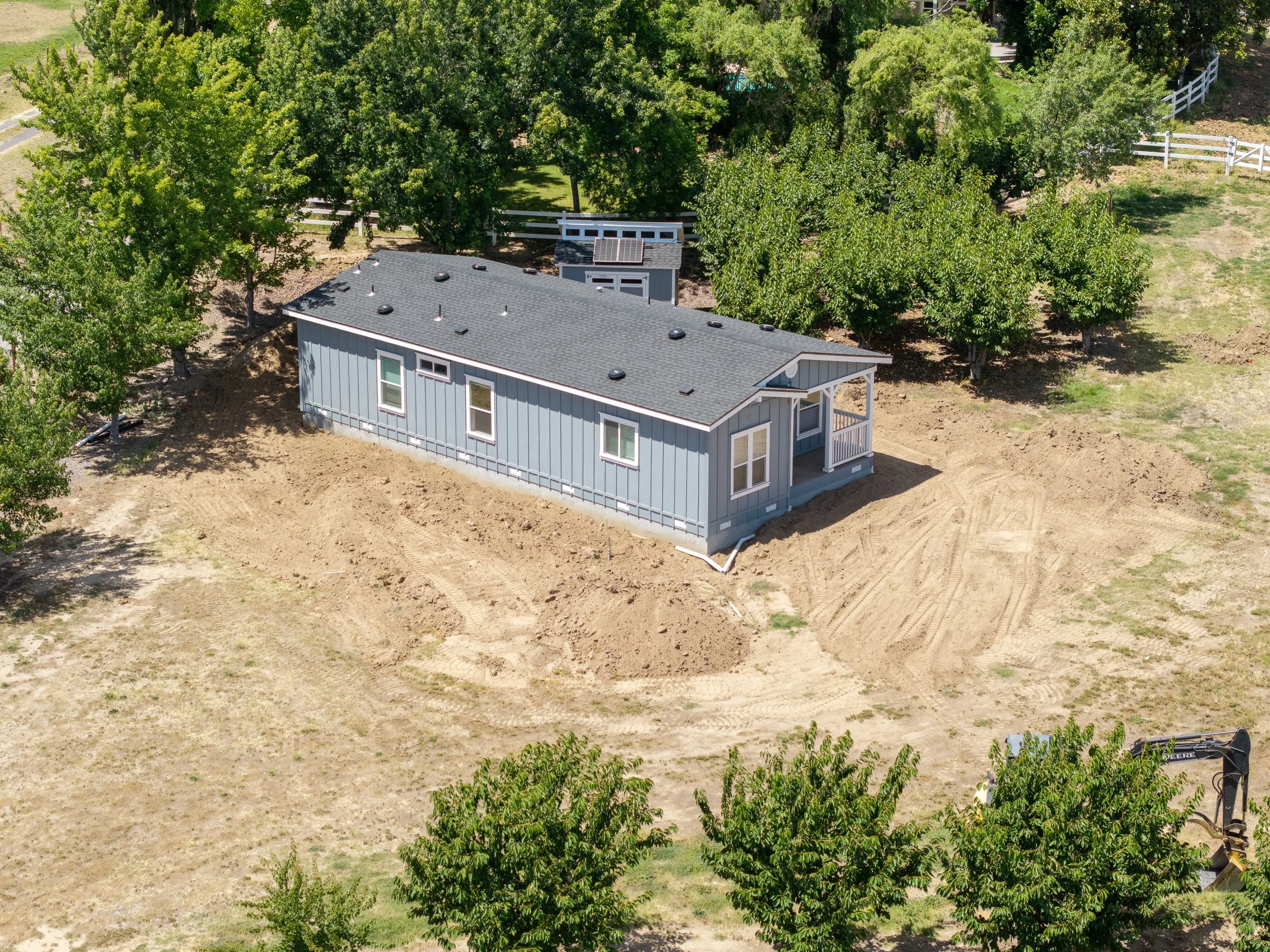 A rooftop house with solar panels surrounded by green trees, fences, and a dirt yard, with construction equipment visible at the bottom right.