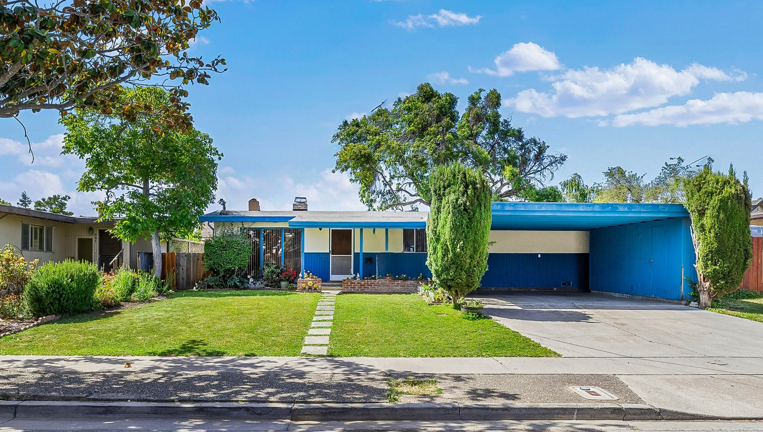 A residential house with a blue rooftop and white walls, surrounded by green trees and a lawn, with a driveway on the right side under a blue sky with clouds.