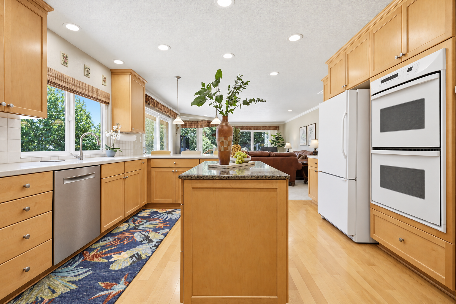Bright kitchen with wooden cabinets, a kitchen island with a decorative vase and fruit, stainless steel dishwasher, white refrigerator, double oven, large windows showing greenery, and a glimpse of a living room with brown couch and art on the walls.