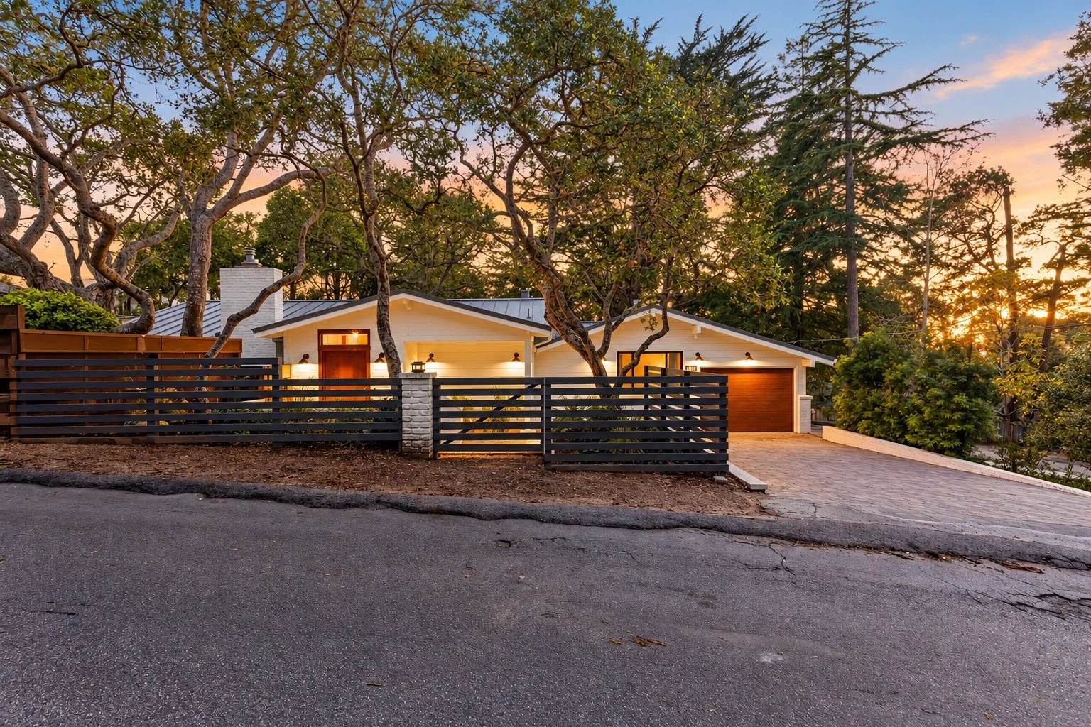 Carmel home at sunset with white siding, a wood garage door, horizontal fencing, and mature trees.