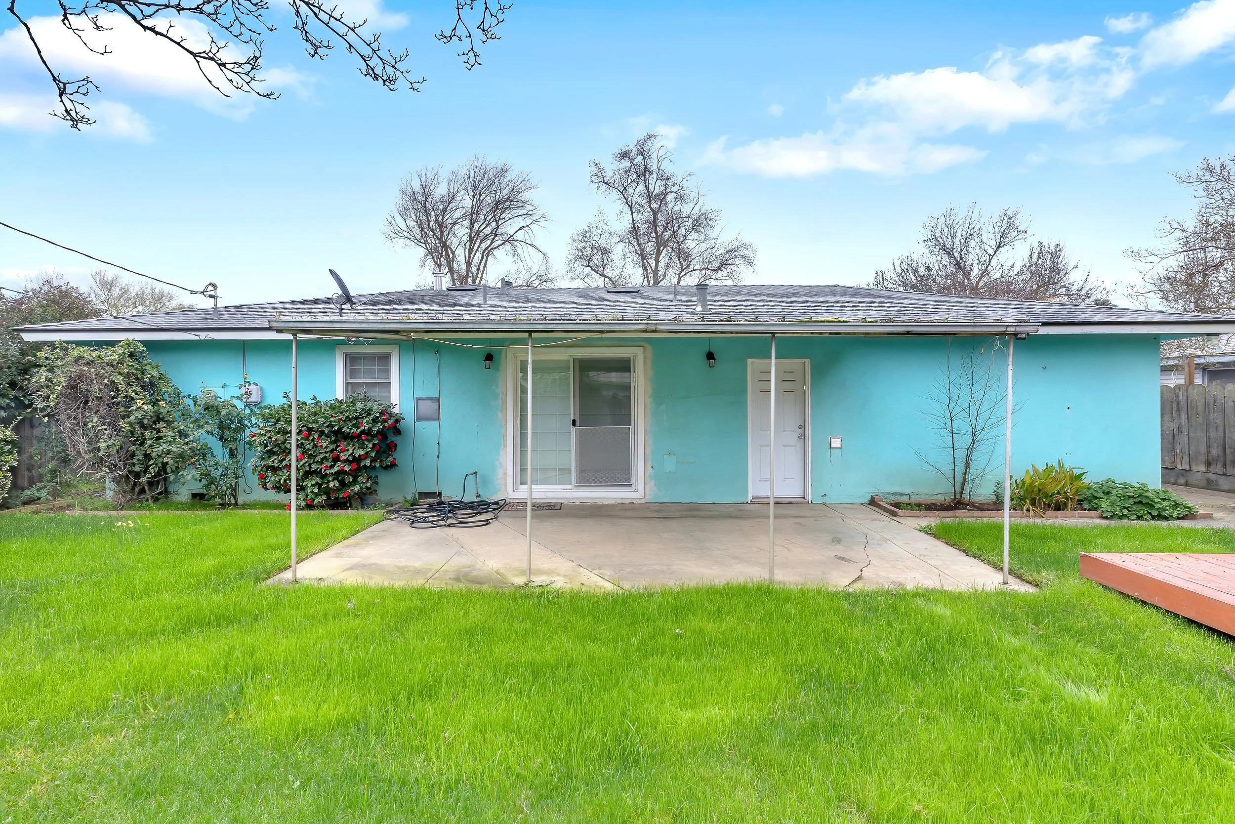 A light blue house with a covered back patio. The yard has a well-maintained green lawn, some bushes, and a small tree. The sky is partly cloudy with a few clouds and leafless trees in the background.