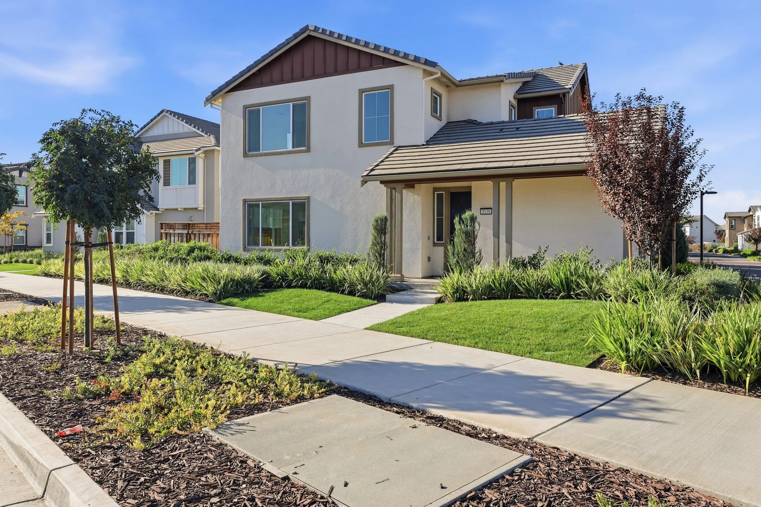 A modern, multi-story residential house with large windows, surrounded by well-maintained grass, trees, and shrubbery, located on a clean sidewalk in a suburban neighborhood.
