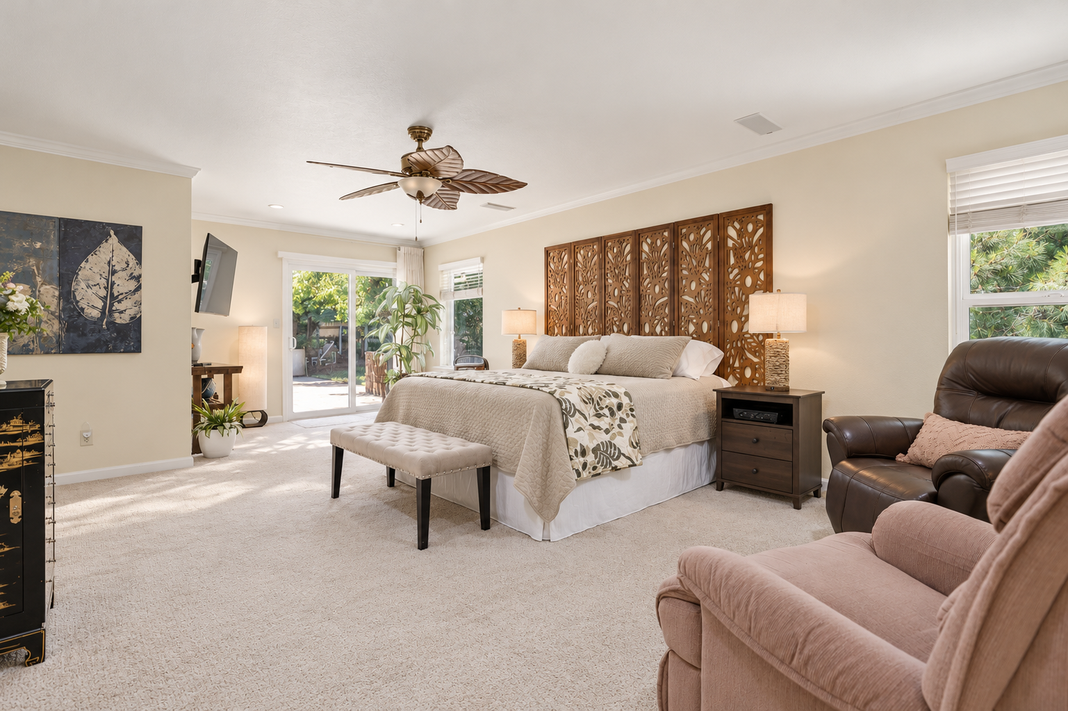 Bright bedroom with a king-sized bed, two bedside lamps, a brown leather armchair, and a sliding glass door leading to an outdoor patio.