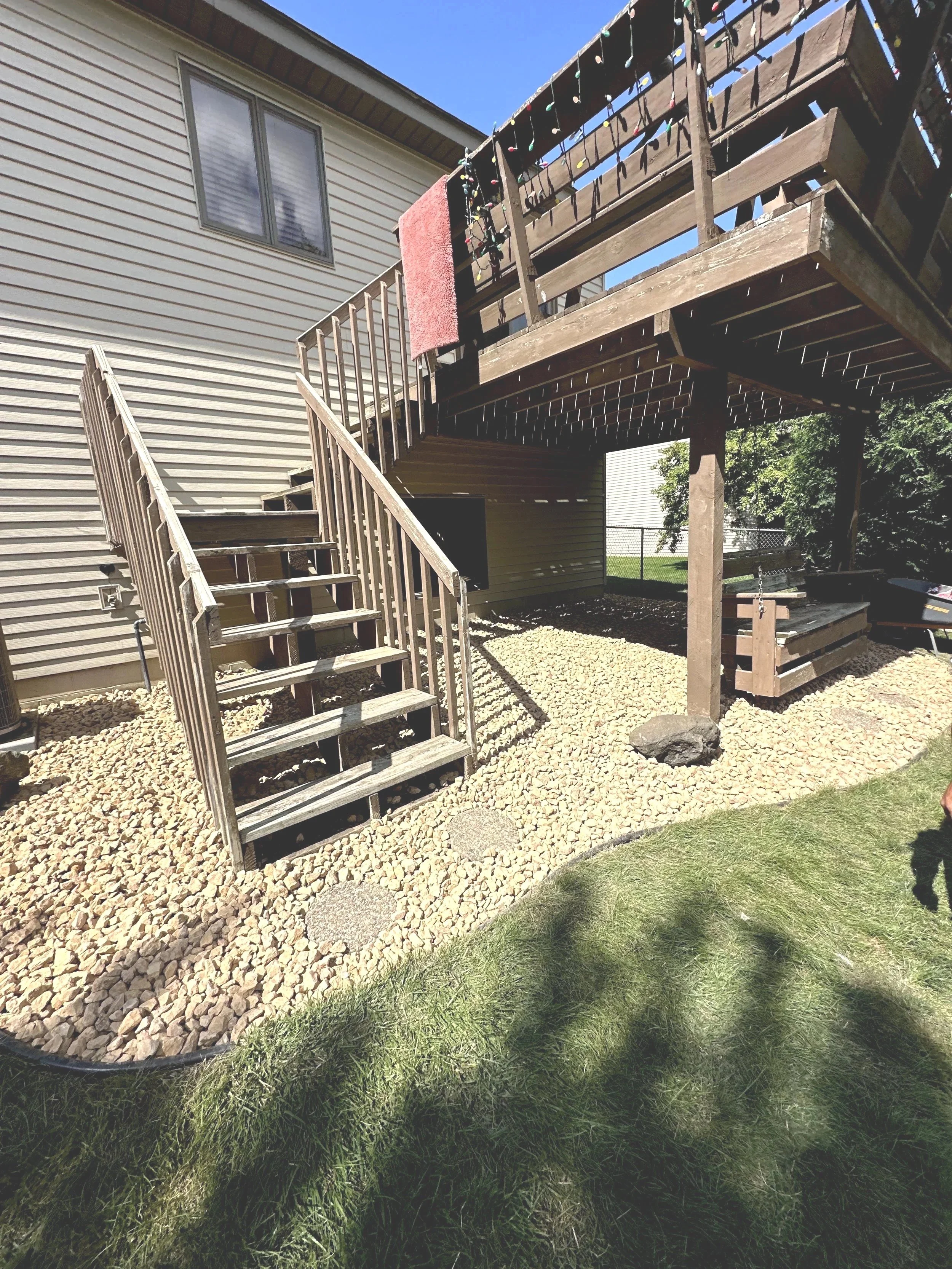 Backyard with a wooden deck and stairs leading down to a gravel area; the deck has Christmas lights and a red towel hanging over the railing; part of a house with horizontal siding is visible.