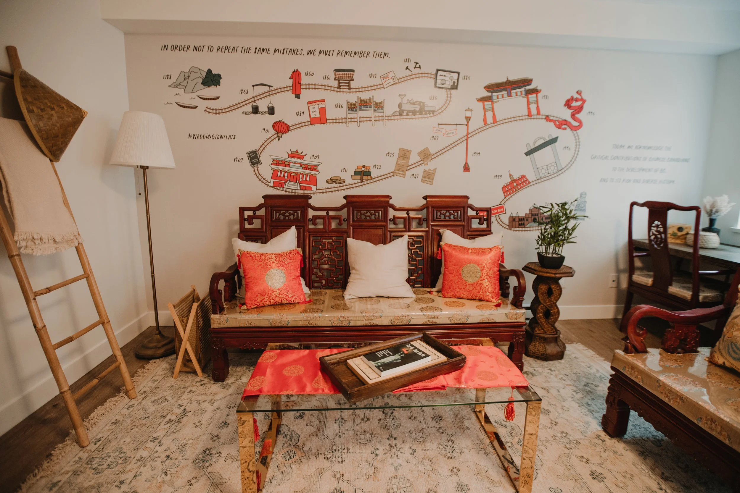 Living room with a carved wooden sofa with two coral-colored accent pillows and one white pillow, a glass-top coffee table with a red cloth and tray, a potted plant on a carved wooden side table, a floor lamp, a bamboo ladder with a beige fabric hanging on it, wall art with a map and quotes, and a wooden dining table with chairs near a window.