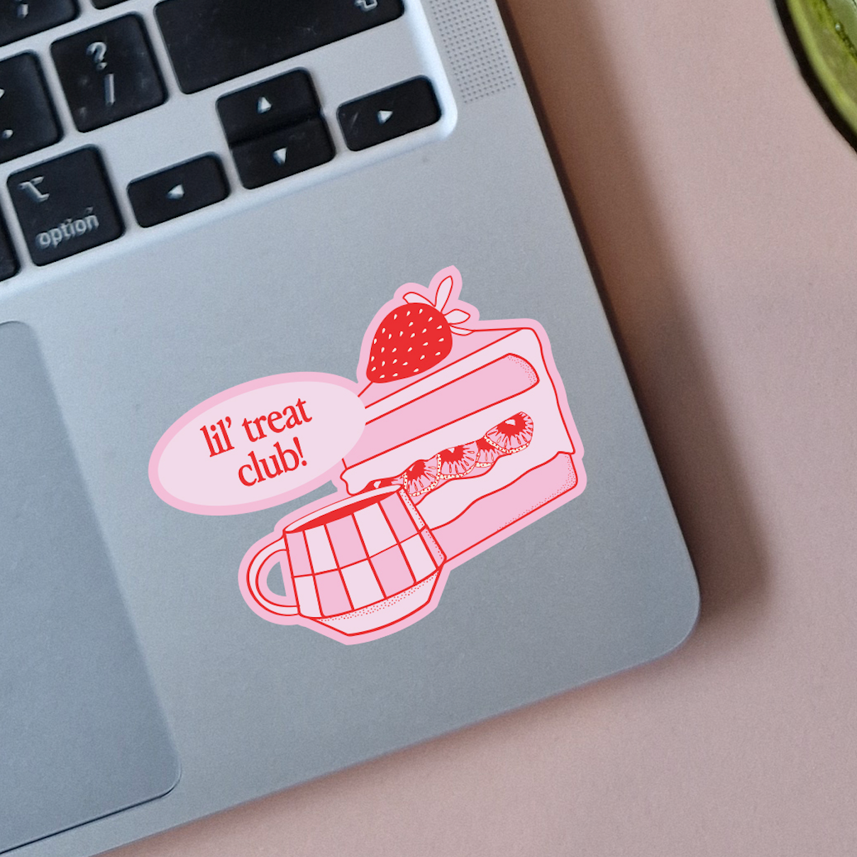 Pink sticker on a laptop featuring a slice of cake with a strawberry on top, strawberries inside the cake, and a fork. Speech bubble from the sticker says 'lil' treat club!'