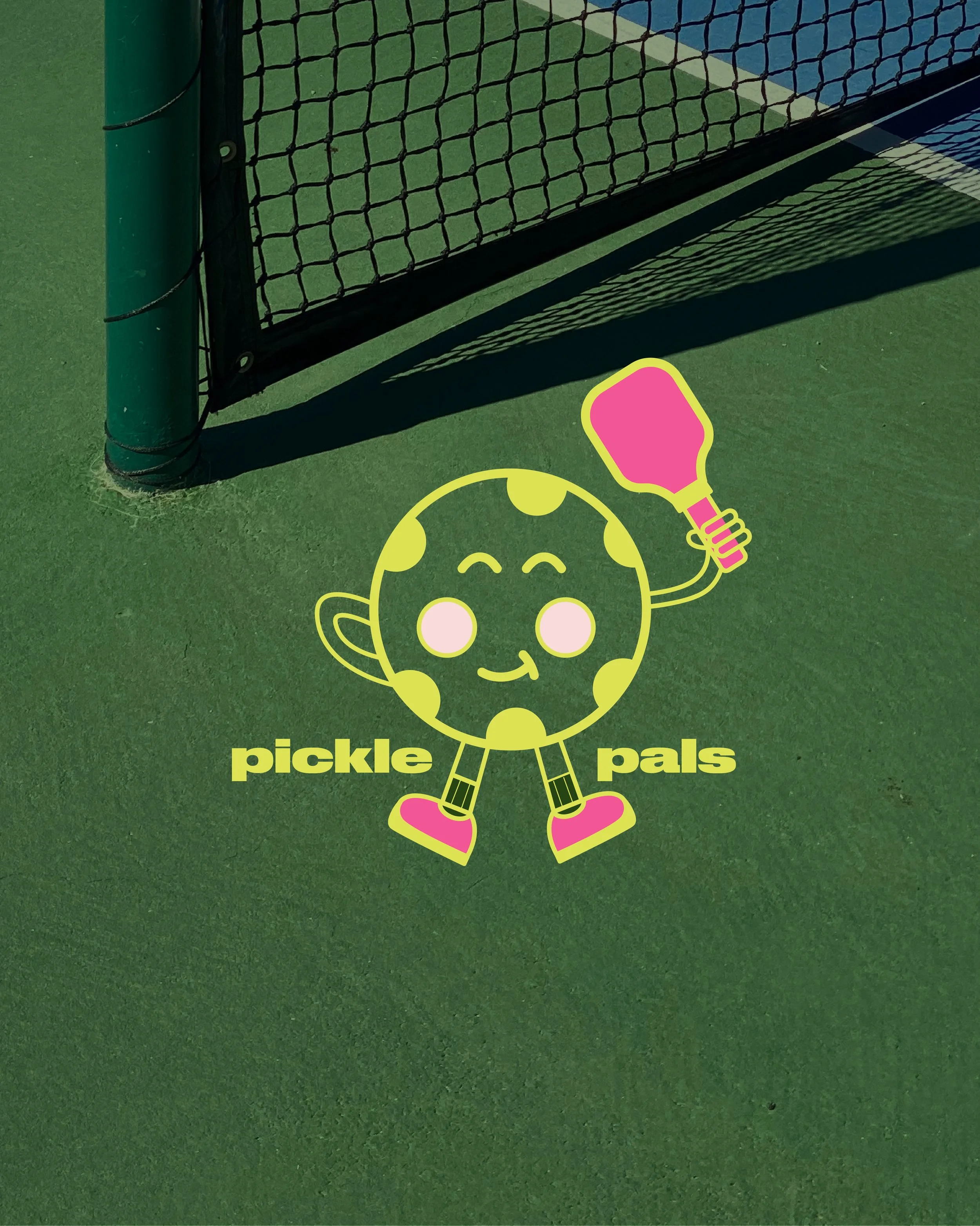 A cartoon graphic of a smiling pickle character holding a pink pickleball paddle. The pickle is wearing pink shoes and has arms and legs, positioned over a green outdoor tennis court near a fence. The words 'pickle pals' are written below the character.