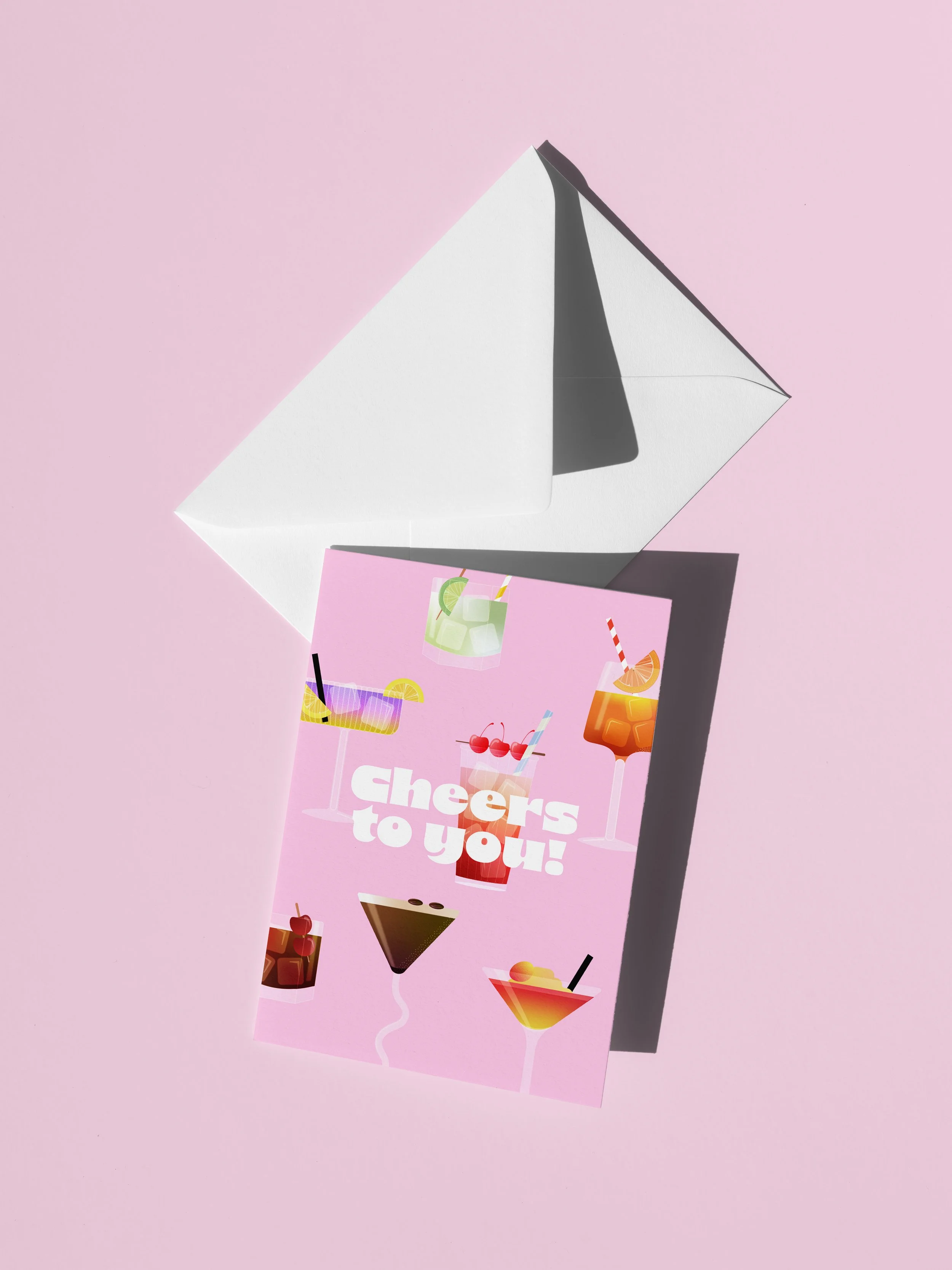 A pink greeting card with colorful cocktail illustrations and the message "Cheers to you!" is placed on pastel pink background, partially leaning against an open white envelope.