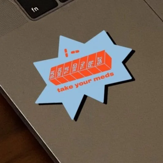 A sticker on a laptop with a comic-style speech bubble design, featuring a red pill organizer and the text 'take your meds' in bold orange letters.