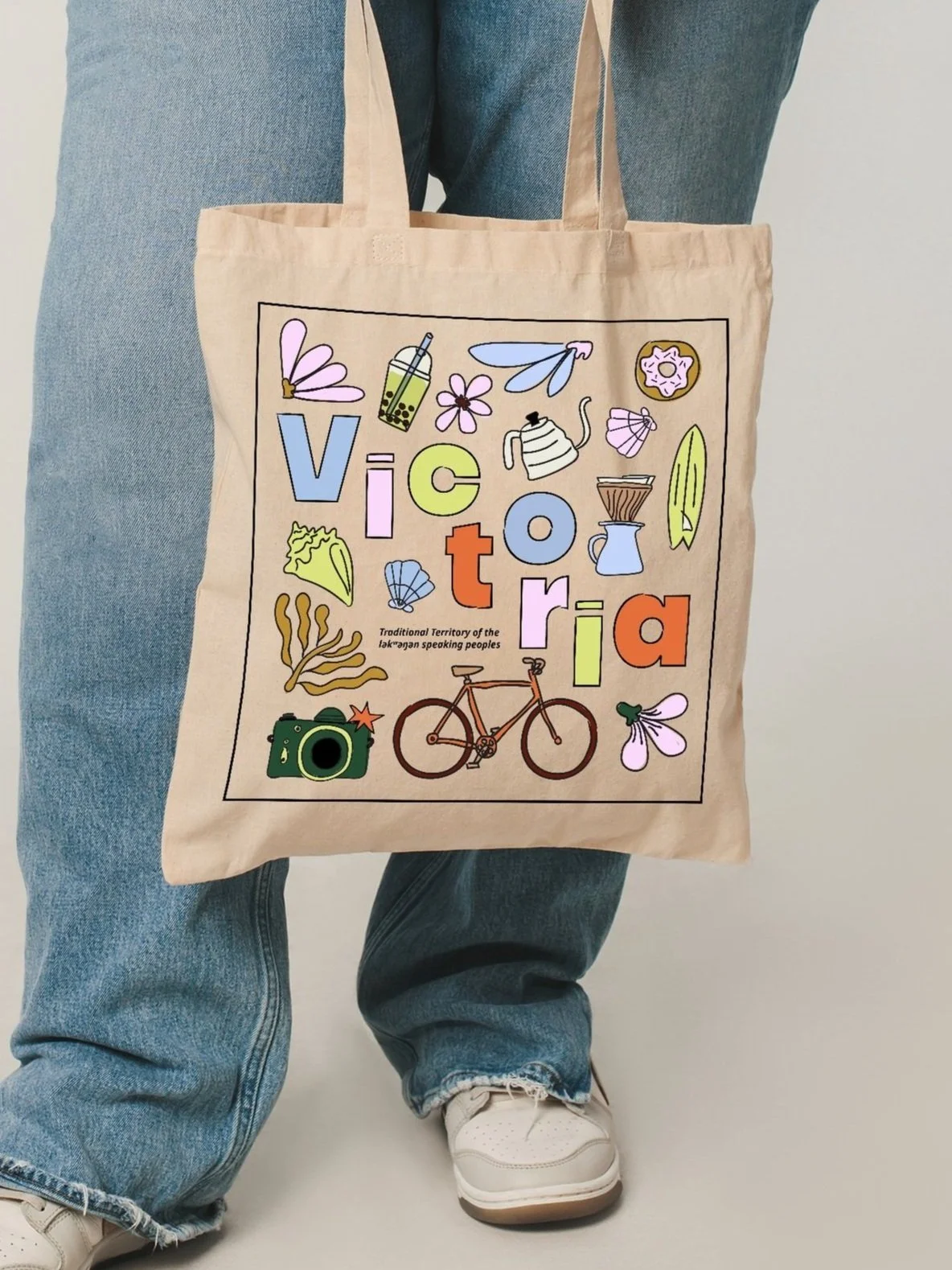 A beige tote bag with colorful illustrations of a bicycle, camera, flowers, teapot, bubble tea, and the word "Victoria" in large, multicolored letters, along with a small text that reads "Traditional Territory of the Lak'wungen speaking peoples."