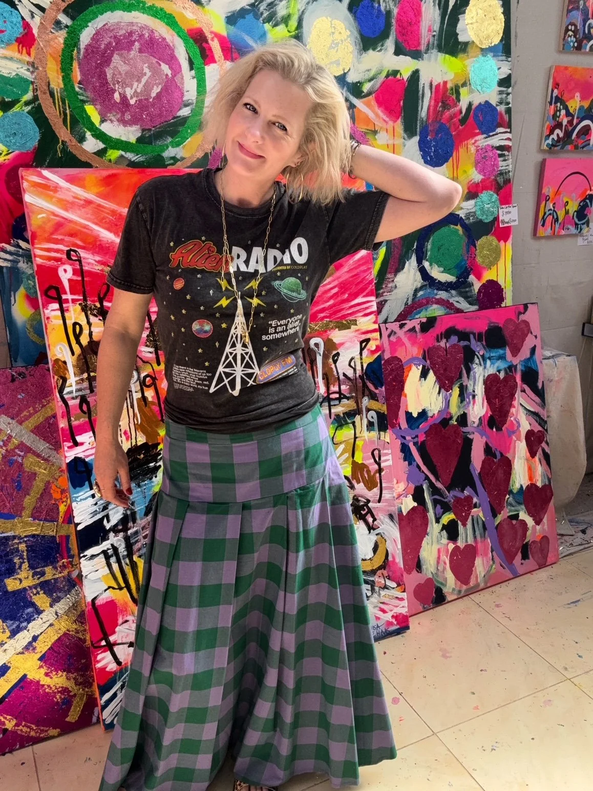 Colourful Australian Artist Louisa de Haas has blonde hair, and is wearing a Coldplay Alien Radio tshirt, a purple and green tartan skirt and bold jewellery. Large, bright abstract paintings are behind her.She is in her Southern Highlands studio.