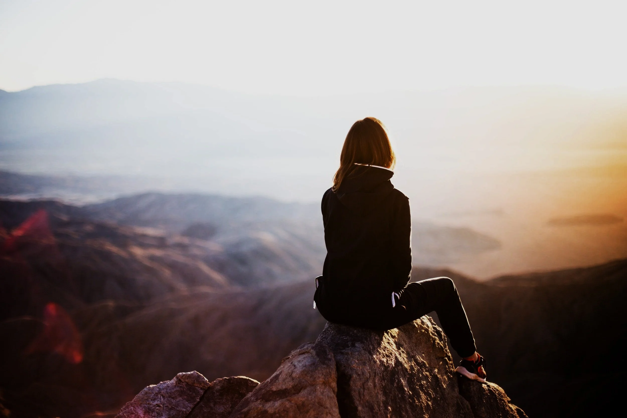 A woman sitting on a rocky ledge overlooking a valley and distant mountains during sunset or sunrise.
