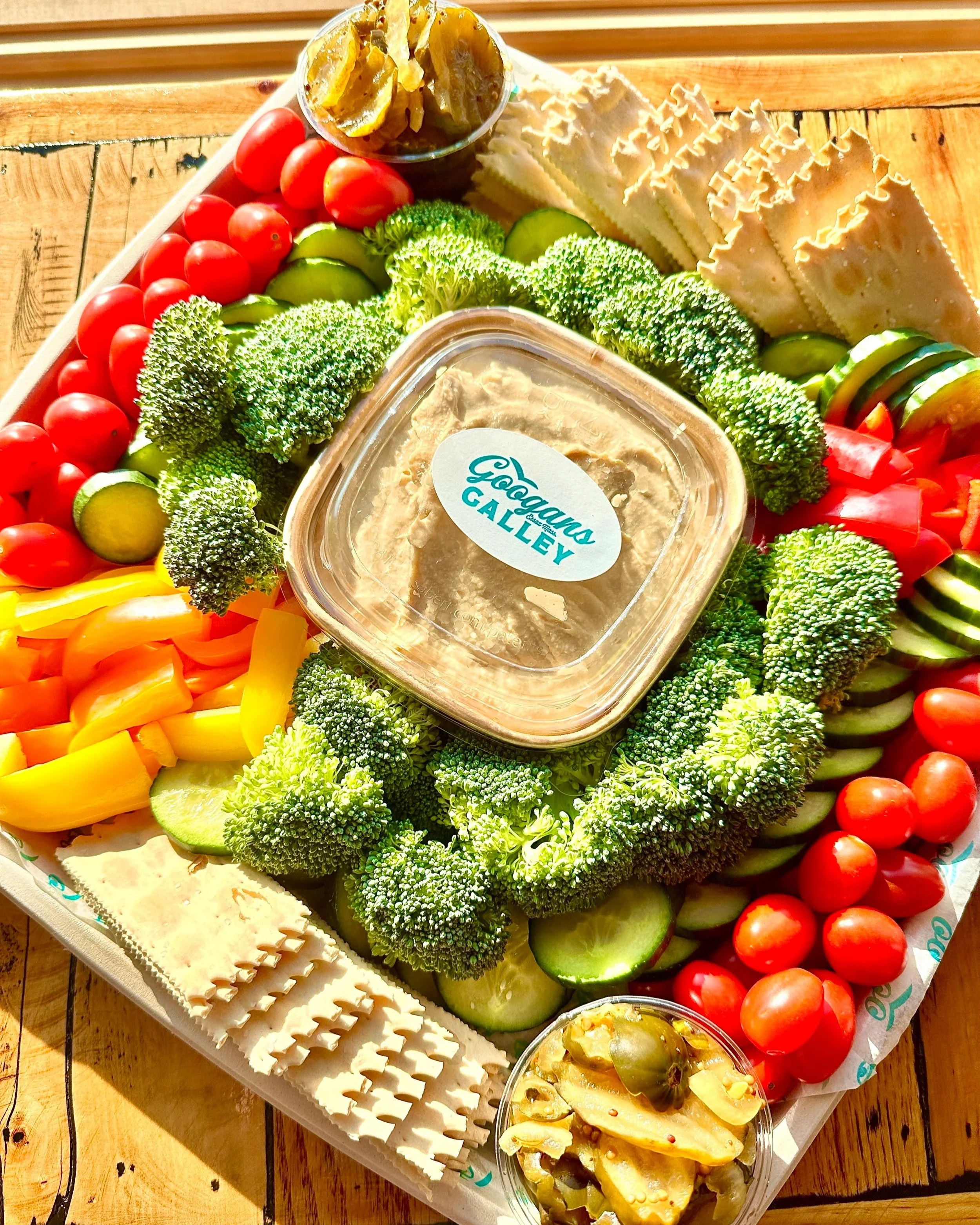 A tray of fresh vegetables and cheeses, including cherry tomatoes, cucumber slices, broccoli, sliced cheese, pickles, and a container of hummus with a label 'Googans Galley'