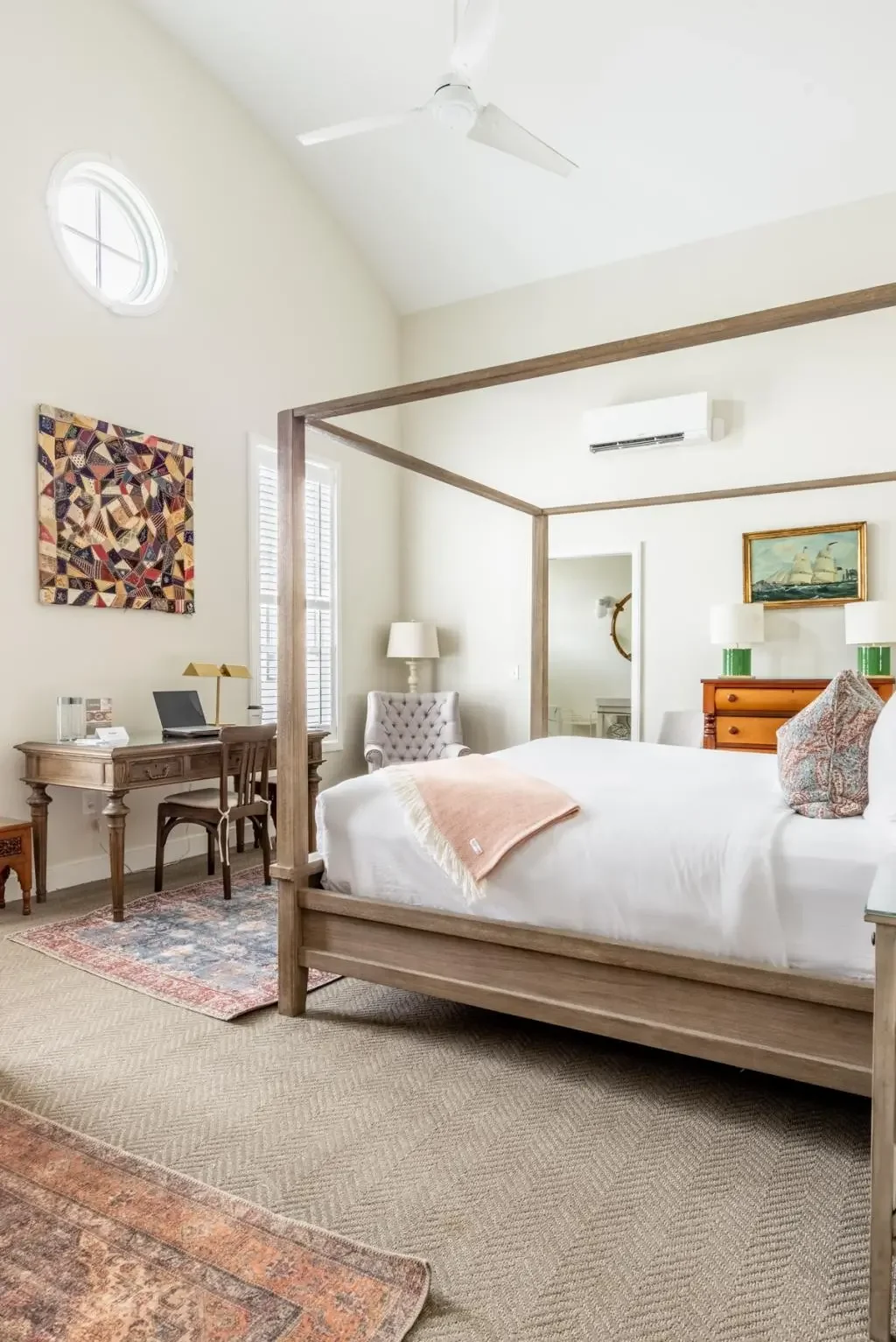 A bright bedroom with a wooden four-poster bed, white bedding, decorative pillows, and a pink throw blanket. The room features large windows with white shutters, a ceiling fan, a wall-mounted air conditioner, a painting of a sailing ship, a vintage wooden dresser with two green lamps, a white armchair, a wooden desk with a chair, and colorful artwork on the wall.